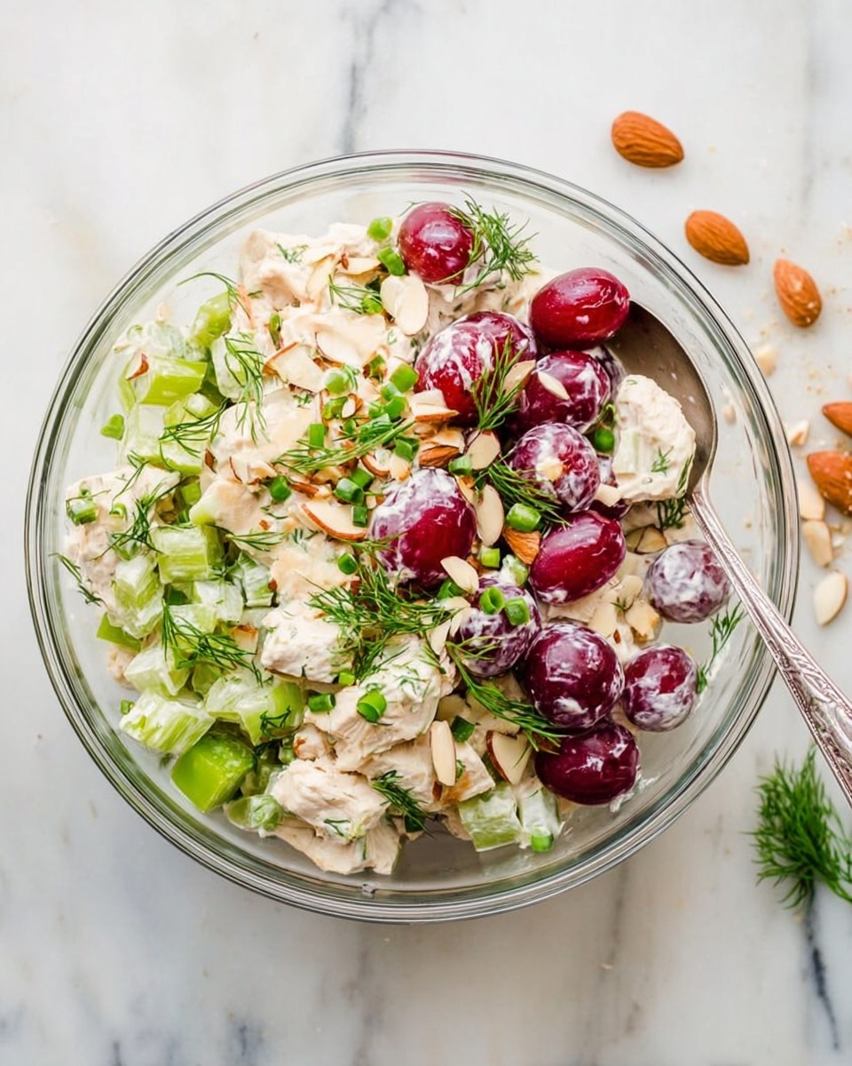 A clear glass bowl filled with a chicken salad sits on a white marbled surface. The salad has three main layers: chunks of white chicken pieces, cut red grapes showing their juicy inside, and chopped green celery mixed evenly throughout. On top, there are small sliced light brown almonds, green herbs like dill, and finely chopped green onions sprinkled. A silver spoon is placed inside the bowl on the right side. Some grapes, almonds, and dill are scattered casually around the bowl for decoration. photo taken with an iphone --ar 4:5 --v 7
