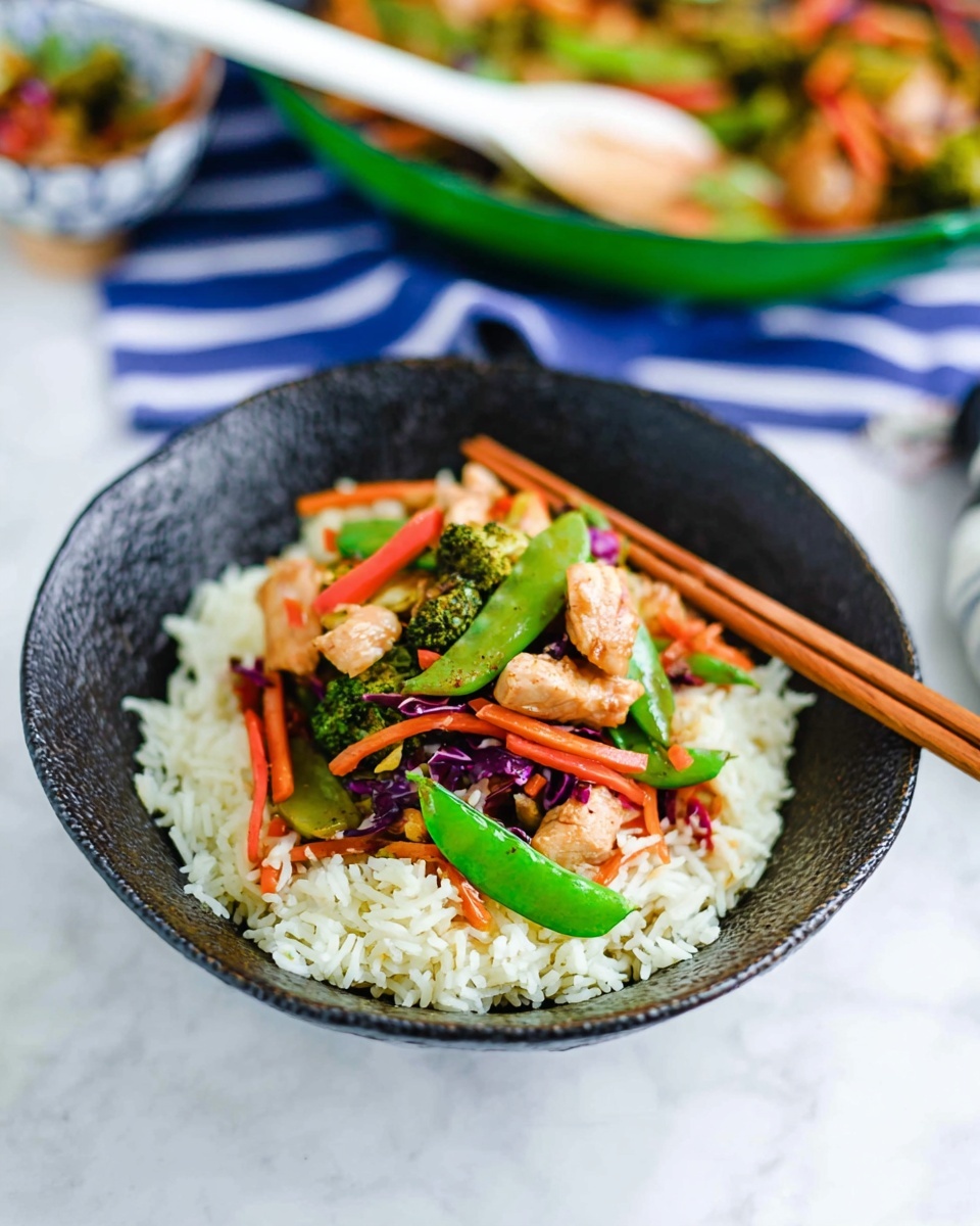 A black textured bowl holds a serving of white rice as the base layer, with a colorful stir-fry mix placed neatly on top. The stir-fry contains vibrant green snap peas, bright orange carrot sticks, small pieces of light brown cooked chicken, purple cabbage, and small broccoli florets, creating a lively mix of colors and textures. The bowl is set on a white marbled textured surface, with a pair of wooden chopsticks resting across the bowl’s edge. In the background, a green pan with more stir-fry and a white spoon inside is slightly out of focus, placed on the same white marbled surface with a blue and white striped cloth nearby. Photo taken with an iphone --ar 4:5 --v 7