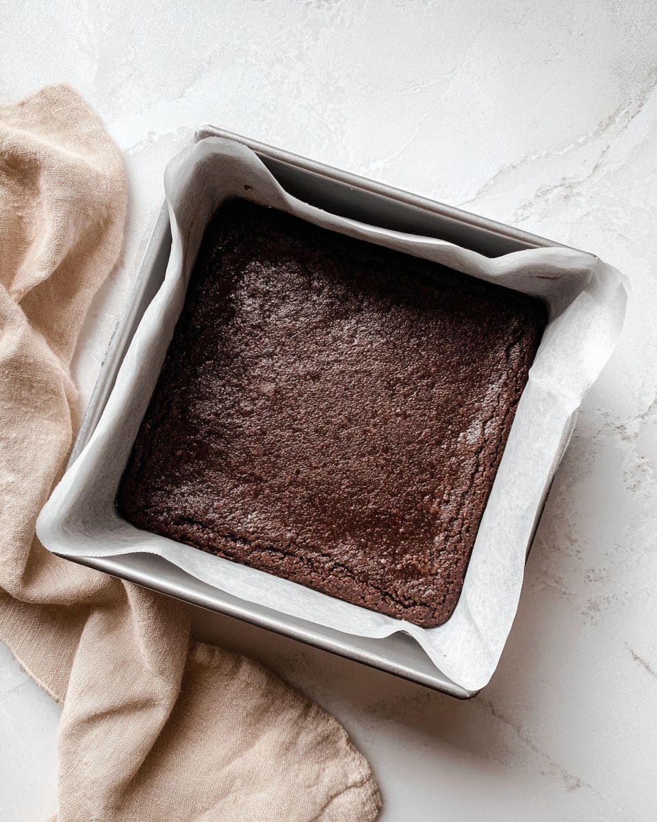 A dark brown, square baked brownie with a rough, slightly cracked top layer is placed inside a silver baking pan lined with white parchment paper. The brownie looks thick and dense, filling the pan evenly. The pan sits on a white marbled textured surface, next to a soft beige cloth draped to the side. photo taken with an iphone --ar 4:5 --v 7