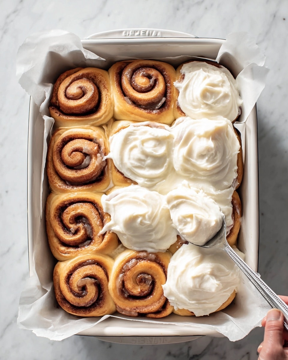 A white baking dish holds nine golden-brown cinnamon rolls arranged in three rows of three, with five of them covered in thick, creamy white frosting that is spreading smoothly over the rolls. The cinnamon rolls show layered spirals of dough with a rich brown cinnamon filling visible inside each swirl. A silver spoon, held by a woman's hand, is spreading more frosting onto the rolls, adding a soft, glossy texture on top. The dish is lined with parchment paper and sits on a white marbled surface. photo taken with an iphone --ar 4:5 --v 7