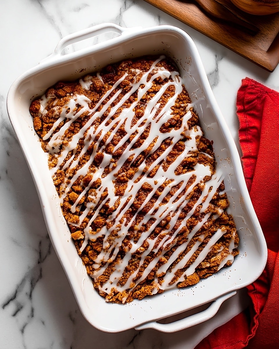 A white rectangular baking dish filled with a crumbly baked dessert that has a golden brown color with uneven texture, topped with a white icing drizzled in thin, irregular lines across the surface. The dish is placed on a white marbled surface, with a folded red cloth visible on the right side and wooden and ceramic kitchen items partially seen at the edges. photo taken with an iphone --ar 4:5 --v 7