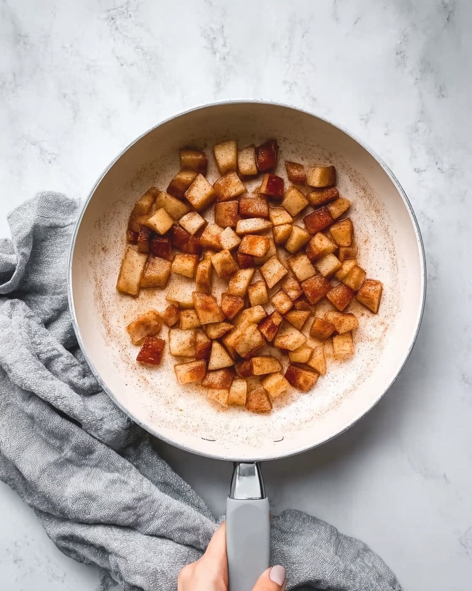 A white pan with a gray handle is placed on a white marbled surface, filled with small, lightly browned cubed pieces of apple scattered evenly inside. A woman's hand gently holds the pan handle, and a soft gray cloth lies near the pan on the surface. The overall colors show warm caramel tones from the cooked apple pieces against the clean white pan and marbled background. Photo taken with an iphone --ar 4:5 --v 7