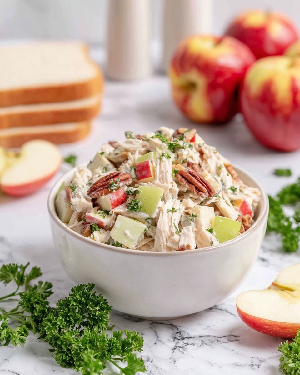 A white ceramic bowl filled with a salad made of shredded light beige chicken, small cubes of red and yellow apple, and sliced pale green celery, all mixed with chopped green parsley and some visible brown pecans. The salad has a creamy texture and is slightly piled over the rim of the bowl. Around the bowl on a white marbled surface, there are sprigs of fresh green parsley, a few celery slices, small apple pieces, and three whole red apples with yellow streaks in the background. There are also two stacked slices of white bread and two white and beige salt and pepper shakers blurred behind the bowl. Photo taken with an iphone --ar 4:5 --v 7