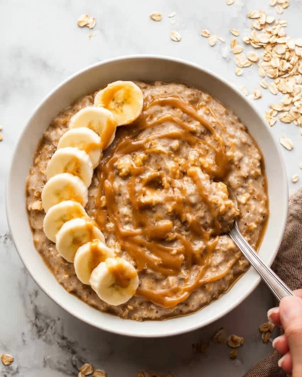 The image shows a white bowl filled with thick oatmeal that has a light brown color with visible oats and swirled caramel sauce on top. One side of the bowl is decorated with five banana slices arranged in a neat row. The bowl is placed on a white marbled surface with some scattered oats and a woman’s hand holding a spoon nearby. Photo taken with an iphone --ar 4:5 --v 7