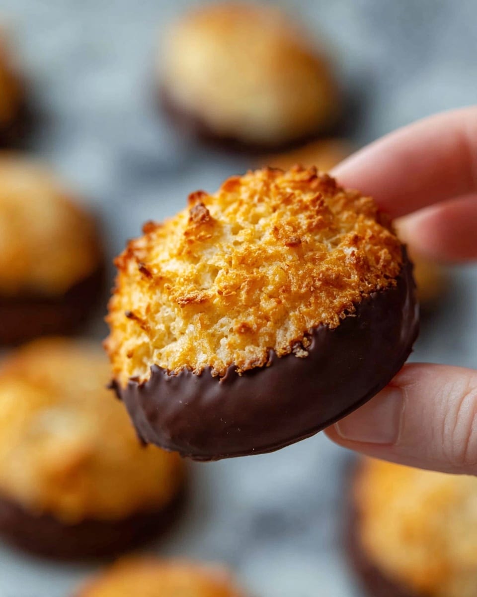 A close-up shows a round, golden brown cookie with a rough, crumbly texture on top, held between a woman's thumb and finger. The bottom half of the cookie is dipped in dark brown chocolate, creating a smooth, shiny layer beneath the bumpy top. In the blurred background, more similar cookies rest on a white marbled surface, all having the same two-tone look with crispy tops and chocolate bottoms. Photo taken with an iphone --ar 4:5 --v 7