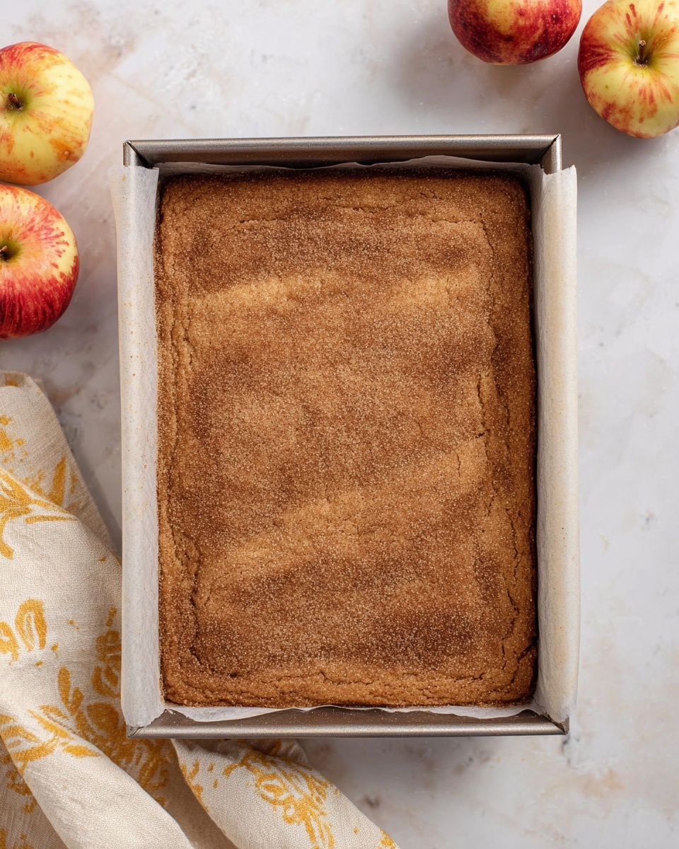 A rectangular brown baked cake with a light dusting of cinnamon sugar on top sits inside a metal baking pan lined with parchment paper, placed on a white marbled surface. The cake has an even, slightly cracked texture and looks soft and moist. Around the pan are two apples with red and yellow patches at the top left, and a cream-colored cloth with a yellow pattern is casually draped at the bottom left corner. The overall setting is simple and clean. photo taken with an iphone --ar 4:5 --v 7