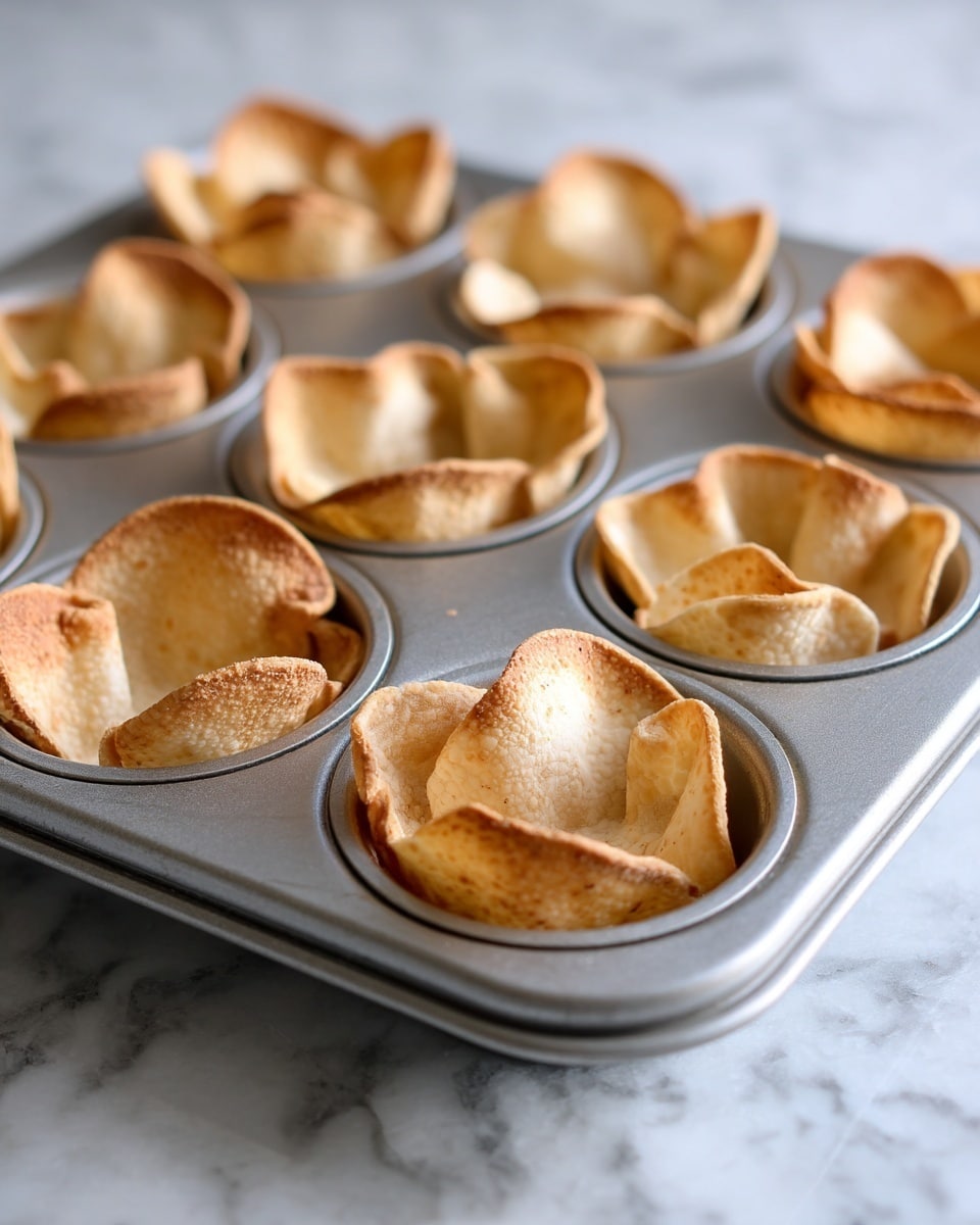 A metal muffin tray filled with round pieces of light brown tortilla shells, each one gently pressed into the cups of the tray to form a shallow bowl shape with small folds and slight bends at the edges, showing some darker toasted spots. The tray sits on a white marbled surface, and the shells look soft and thin with a smooth texture. Photo taken with an iphone --ar 4:5 --v 7