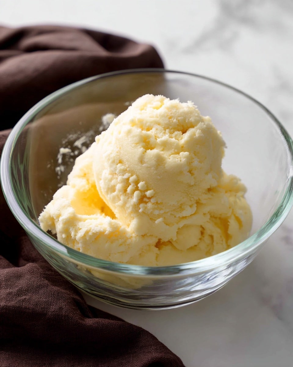 A clear glass mixing bowl holds a single, large scoop of light yellow, soft, and smooth dough with a slightly crumbly texture. The bowl is placed on a white marbled surface, and a dark brown cloth napkin lies nearby on the bottom left side. The dough sits loosely, showing some small peaks and gentle folds inside the bowl. Photo taken with an iphone --ar 4:5 --v 7