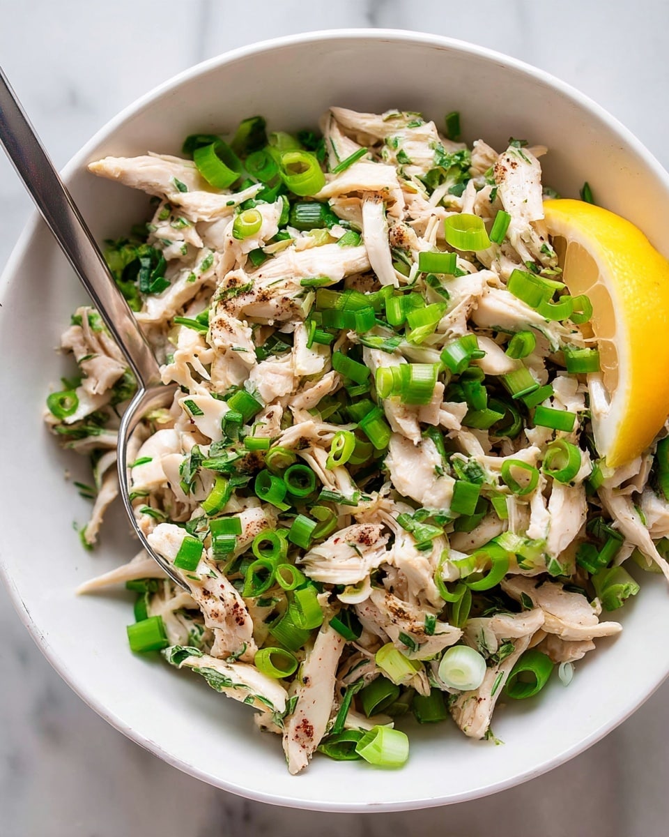 A white bowl filled with shredded white chicken mixed with light creamy sauce, sprinkled with finely chopped green onions and fresh herbs, creating a mix of green and white colors with small dark specks of pepper. Two bright yellow lemon wedges rest on one side of the bowl, adding a fresh and vibrant touch. The bowl is placed on a white marbled surface. photo taken with an iphone --ar 4:5 --v 7