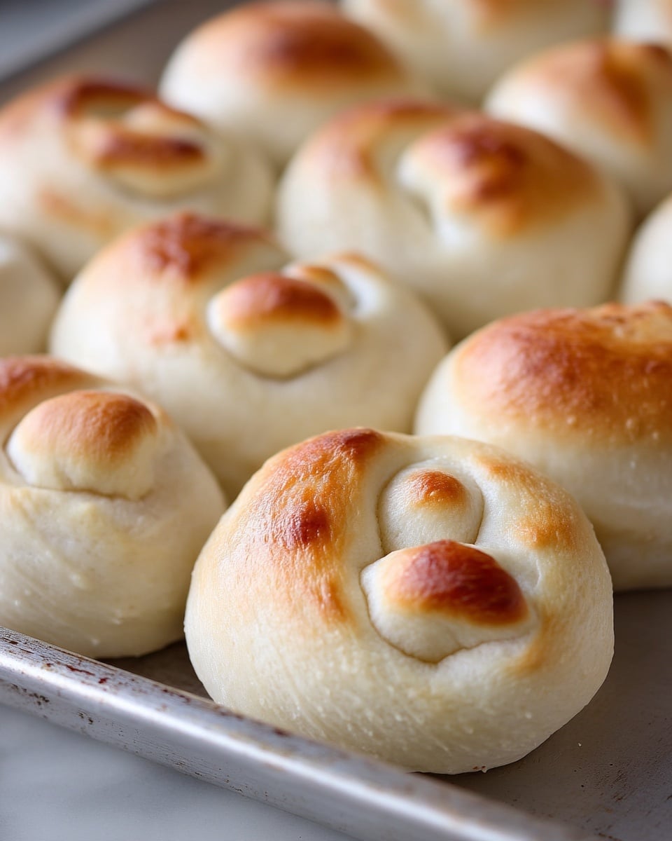 The image shows several freshly baked rolls arranged closely together on a metal baking tray. Each roll has a light golden-brown top with a small round dough piece attached on one side, creating a layered effect with smooth, slightly shiny, and soft-looking textures. The rolls are mostly pale with soft brown highlights from baking, and their shapes are rounded and compact, giving a cozy, fresh-baked appearance. The white marbled texture is visible faintly at the edges under the tray. photo taken with an iphone --ar 4:5 --v 7