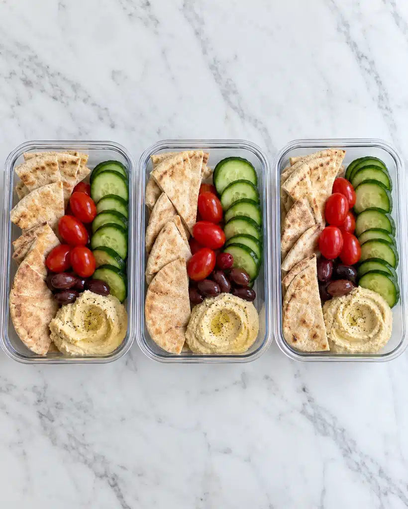 The Hummus Lunch Box Recipe 5 The image shows three clear rectangular meal prep containers placed side by side on a white marbled surface. Each container is divided into two equal sections: the top half contains three layers – sliced green cucumber rounds on the right side, bright red grape tomatoes clustered in the middle, and folded beige pita bread pieces on the left side. The bottom half is split into two parts: the left section holds a small pile of dark brown olives and the right section has a smooth, light beige hummus spread. The colors are fresh and natural, creating a neat and healthy visual appeal. photo taken with an iphone --ar 4:5 --v 7