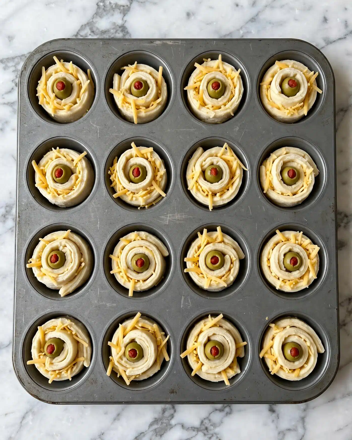 Spooky Eyeball Olive Puffs Recipe 5 A grey metal cupcake tray filled with twelve small rolled dough snacks, each made of one visible layer of pale dough spiraled into a rose shape. The rolls have a green olive with a red center placed in the middle of each swirl. Around the olives, shredded yellow cheese is scattered on top and within the dough spiral layers. The tray rests on a surface with a white marbled texture. photo taken with an iphone --ar 4:5 --v 7