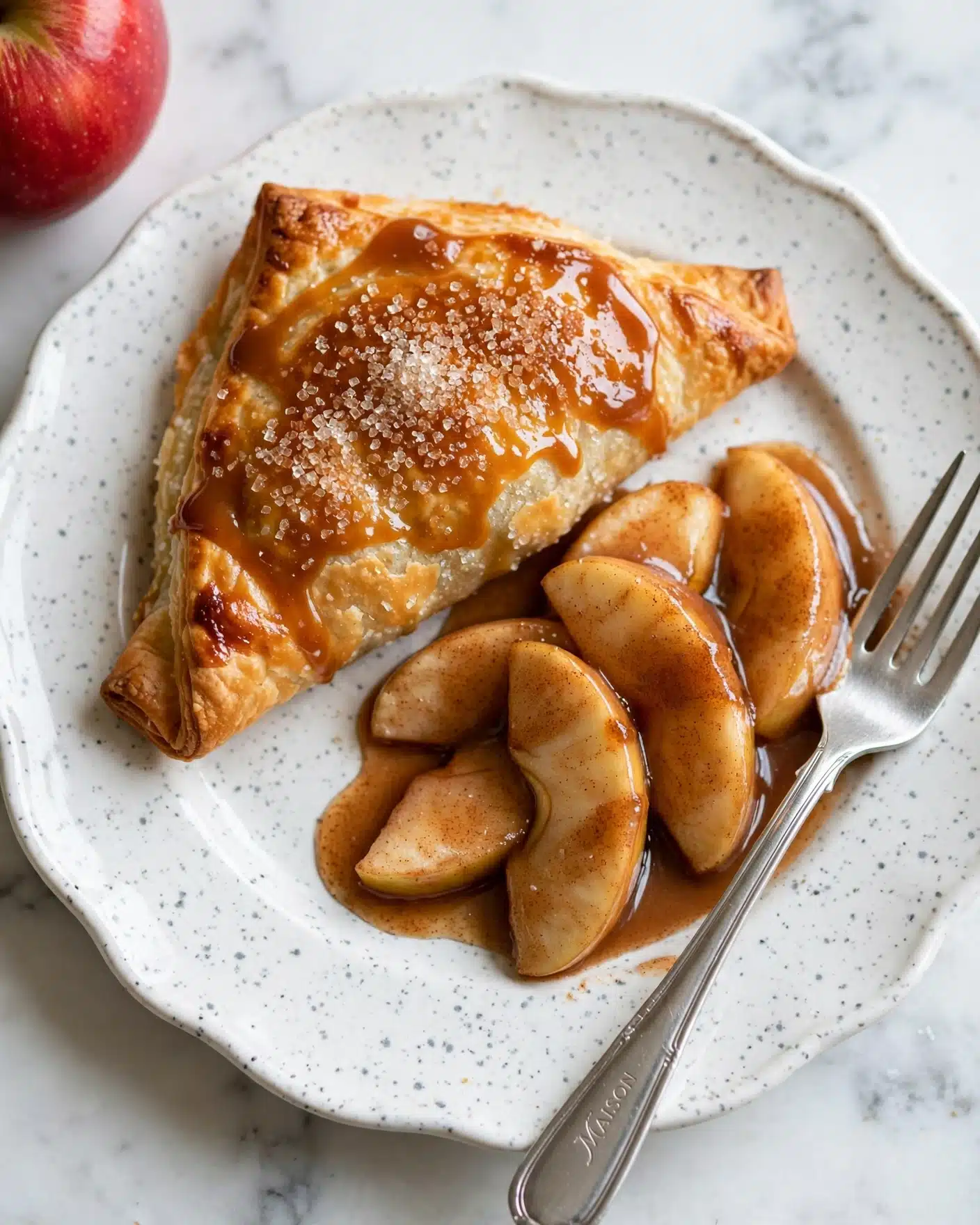 Easy Apple Galette Recipe 5 On a white plate with a speckled pattern and scalloped edges, there is a folded pastry with a golden-brown crust topped with coarse sugar crystals and a shiny caramel glaze. Next to it are soft, sliced apple pieces covered in cinnamon and syrup that has pooled slightly around them. A silver fork rests diagonally across the plate beside the apple slices, its handle engraved with text. The plate sits on a white marbled surface, and part of a red apple is visible in the top left corner of the image. photo taken with an iphone --ar 4:5 --v 7
