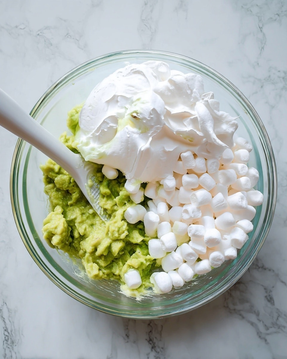 A clear glass bowl sits on a white marbled surface filled with three distinct layers: on the bottom, there is a layer of mashed green avocado with a soft, slightly chunky texture; on one side, a pile of small, white mini marshmallows with a smooth, pillowy look; and on the other side, a fluffy white whipped cream layer with soft peaks. A white spatula rests inside the bowl, touching the avocado layer. photo taken with an iphone --ar 4:5 --v 7