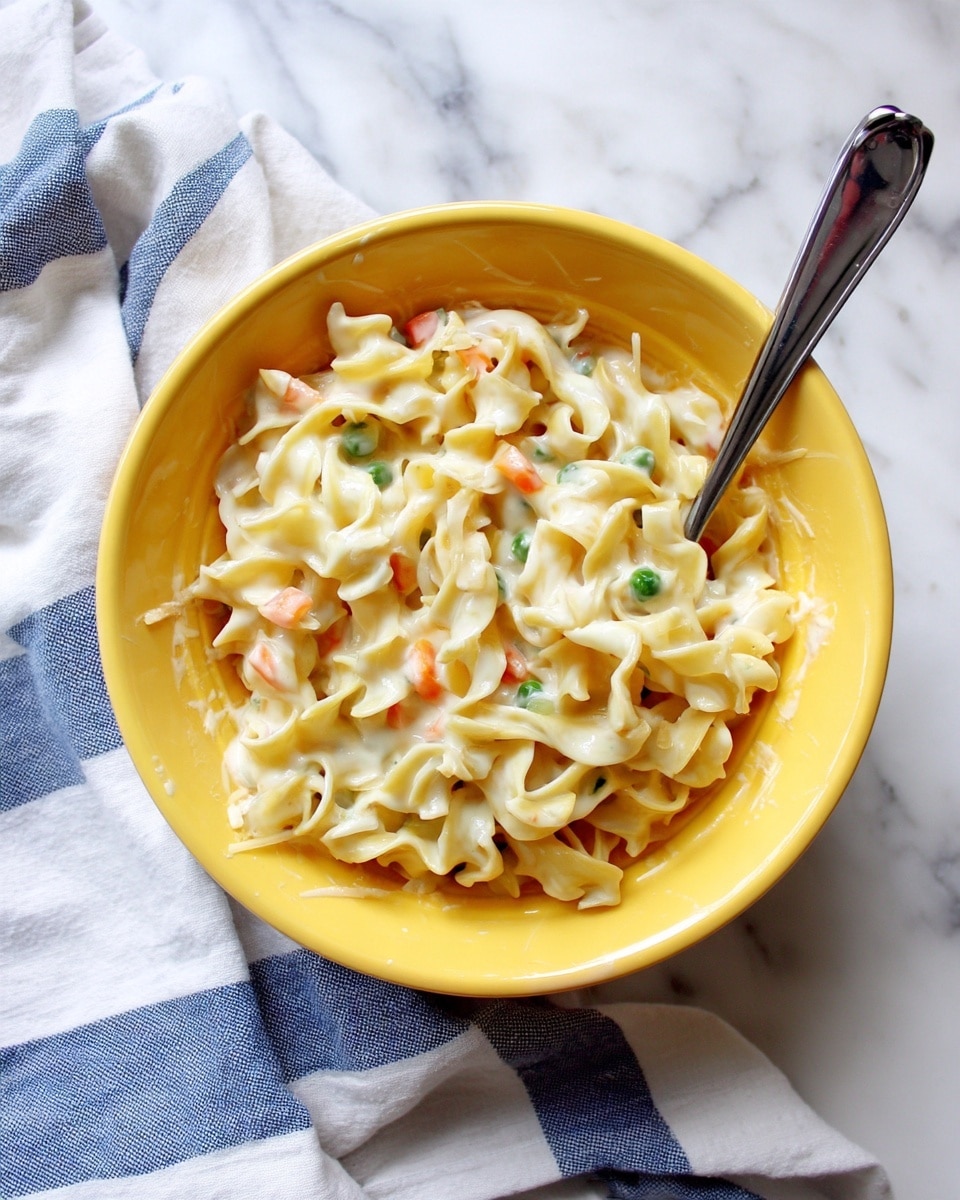 A yellow bowl filled with creamy egg noodles mixed with small pieces of carrot and green peas. The noodles are soft and curly, coated in a thick white sauce with bits of shredded cheese visible. A metal fork rests inside the bowl, partially covered by the noodles. The bowl is placed on a white cloth with blue stripes, all sitting on a white marbled textured surface. photo taken with an iphone --ar 4:5 --v 7