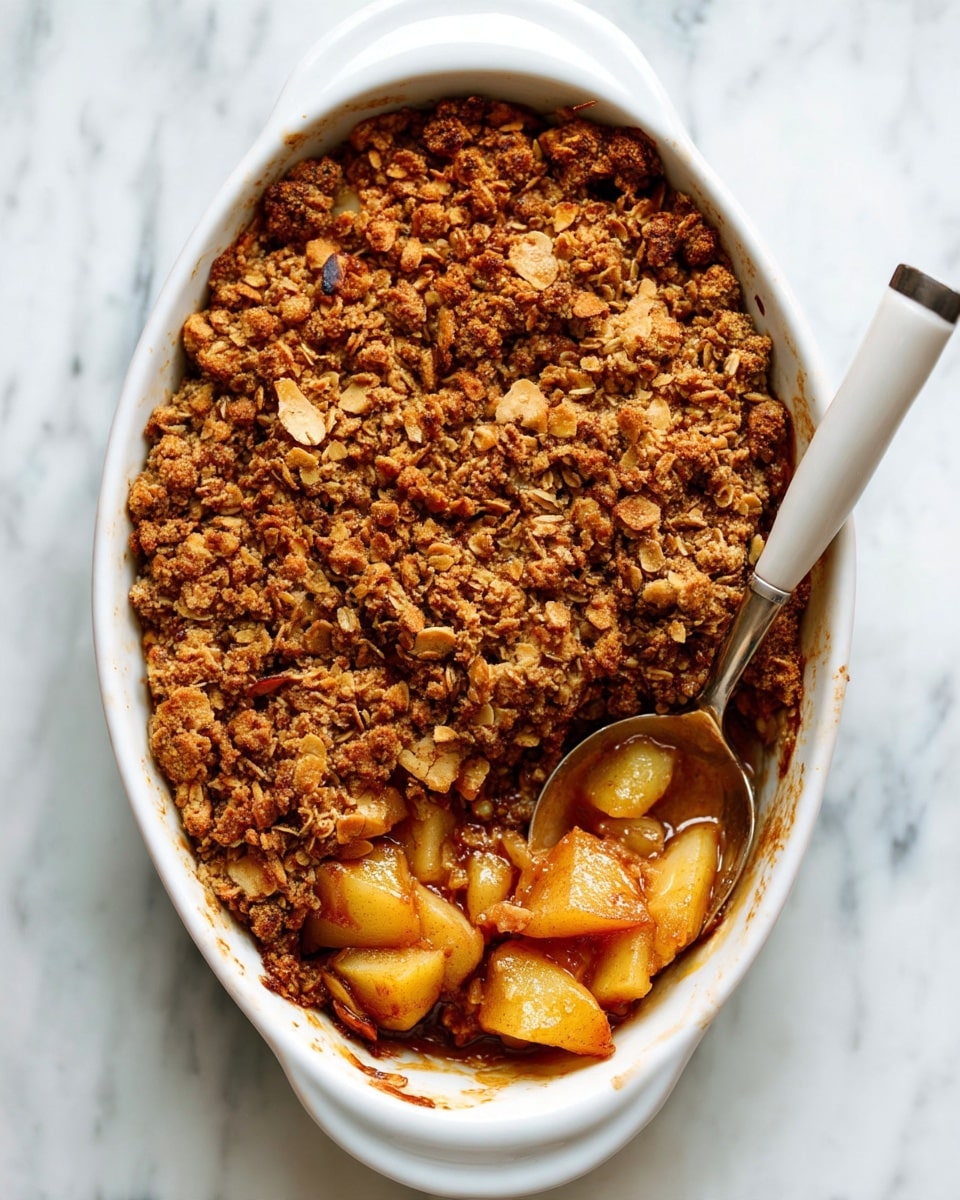 A white oval baking dish filled with a baked fruit crumble, showing two layers: the bottom layer is a mix of soft, light golden chunks of baked fruit with a juicy texture, and the top layer is a thick, crumbly oat and nut mixture in a warm brown color with a coarse texture. A large spoon with a white handle rests inside the dish and scoops some of the fruit and crumble, revealing the glossy fruit juice beneath. The dish is set on a white marbled surface. Photo taken with an iphone --ar 4:5 --v 7