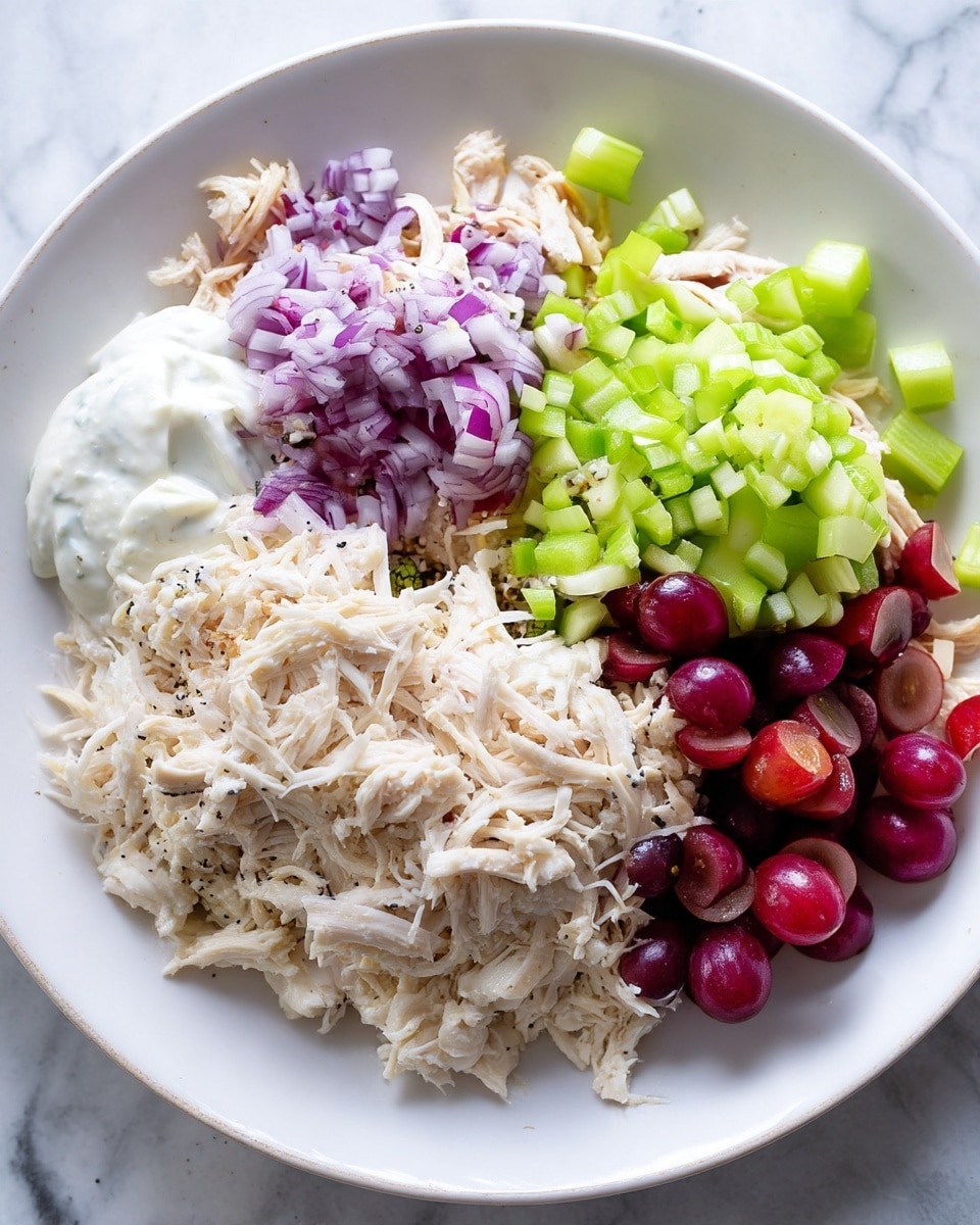 A white plate holds a layered mix of finely shredded pale chicken forming the base layer, topped with small chopped light purple onions placed near the top center. To the right side are bright green, diced celery pieces, while the bottom right features halved dark red grapes showing their juicy interiors. On the left side, there is a creamy dollop of white dressing mixed with a sprinkle of white, black, and brown seeds and spices adding texture. The background and surface are a white marbled texture. Photo taken with an iphone --ar 4:5 --v 7