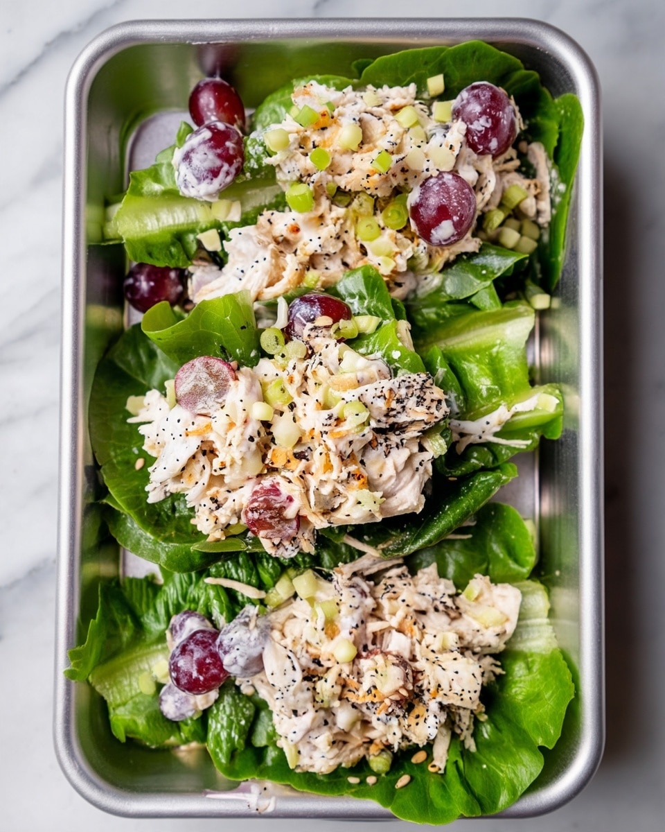 The dish shows three portions of chicken salad placed on fresh green romaine lettuce leaves inside a metal tray. Each portion has shredded white chicken mixed with creamy white dressing, sliced red grapes, and finely chopped green onions scattered on top. The salad is sprinkled with black and white sesame seeds and crispy toasted bits, adding texture and color contrast. The bright green lettuce leaves form a fresh base at the bottom, with the light, creamy salad piled on top. The background is a white marbled texture. photo taken with an iphone --ar 4:5 --v 7