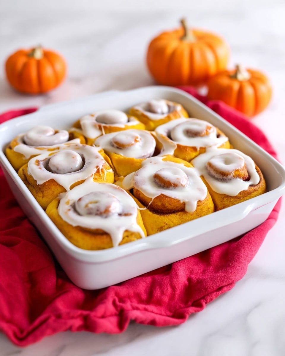 A white baking dish filled with eight pumpkin-colored cinnamon rolls arranged in two rows of four. Each roll has a soft, fluffy texture with a swirl pattern and is generously covered with thick, white creamy icing that slowly drapes over the edges, contrasting with the warm orange dough underneath. The baking dish sits on a bright red cloth against a backdrop of small, bright orange pumpkins and a soft wooden surface beneath, all on a white marbled texture. photo taken with an iphone --ar 4:5 --v 7