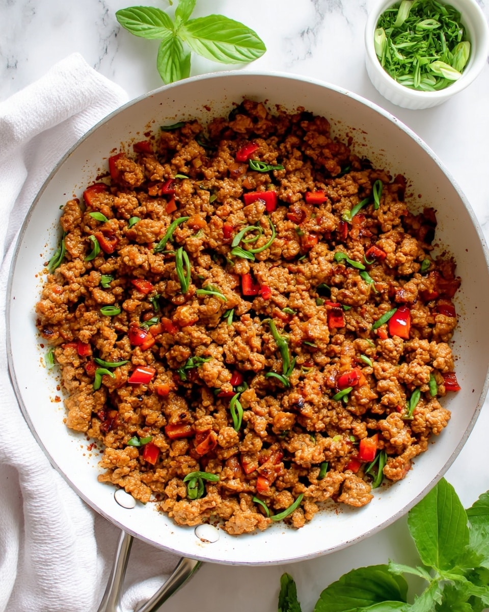 A close-up top view of cooked ground meat mixed with small red bell pepper pieces and scattered green herbs in a white pan. The meat is brown with a slightly crumbly texture, and the red peppers add bright red spots all over. The green herbs are thin strips and scattered lightly on top. Around the white pan, there is a white cloth on the left, a small white bowl with chopped herbs on the top right, and green leaves near the bottom right on a white marbled surface. photo taken with an iphone --ar 4:5 --v 7