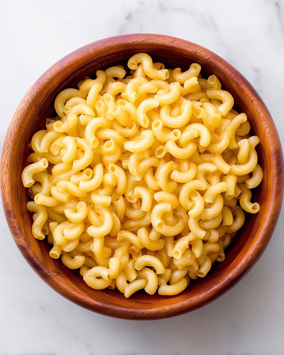 The image shows a close-up view of a round bowl filled with cooked elbow macaroni pasta. The pasta pieces are curved tubes, pale yellow in color, and have a smooth texture with slightly visible ridges. The bowl appears to be wooden with a rich brown color and natural wood grain texture on the inside and outside edges. The background is a white marbled texture that contrasts with the bowl and pasta, making the yellow macaroni stand out. The pasta fills the bowl completely, creating a dense and uniform layer. photo taken with an iphone --ar 4:5 --v 7