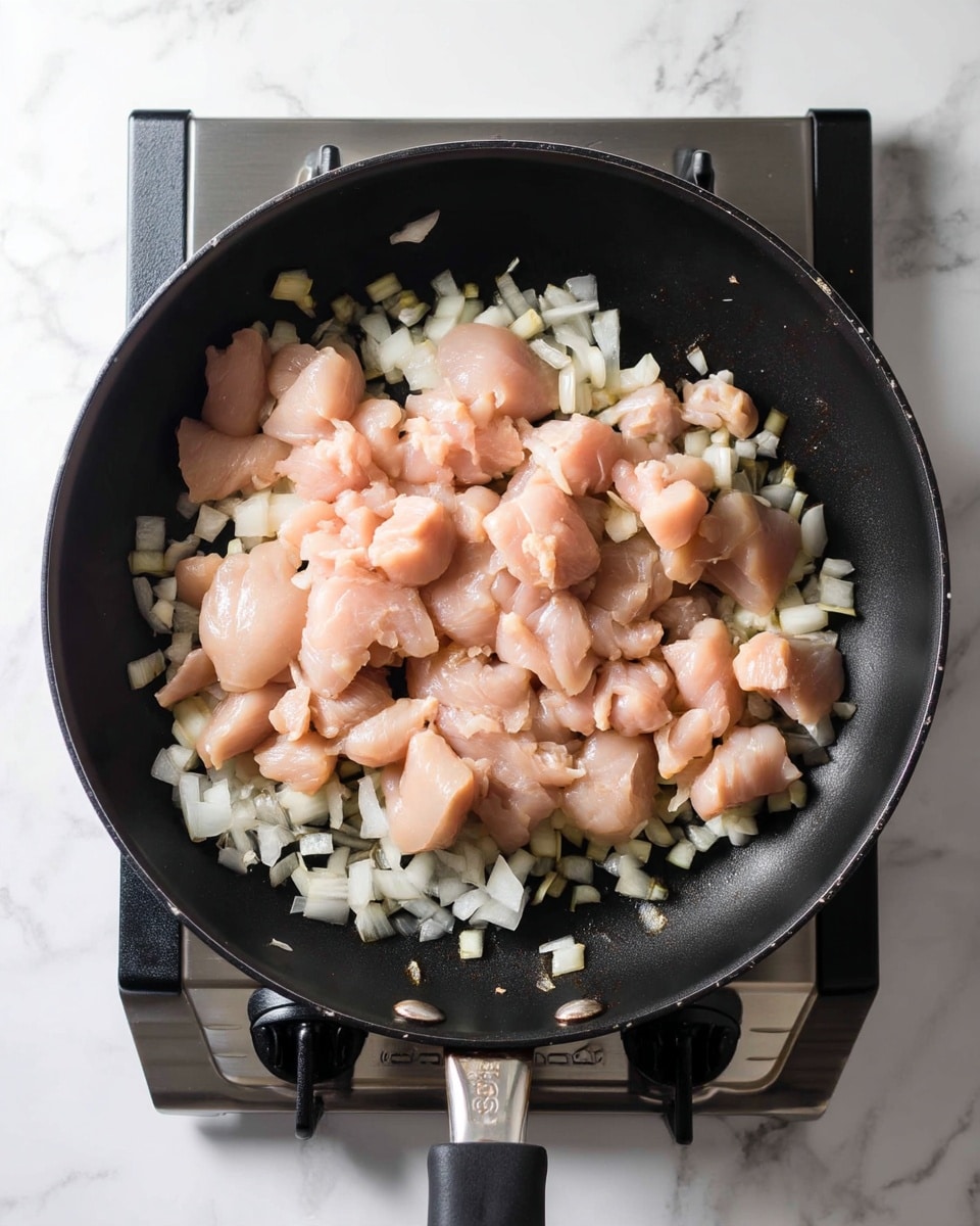 A black frying pan filled with small pieces of raw light pink chicken spread evenly on top of a layer of white chopped onions. The onions have a slightly translucent texture, showing they have started cooking. The pan sits on a stove burner with a simple black metal grate visible under it. The background is a white marbled texture. photo taken with an iphone --ar 4:5 --v 7