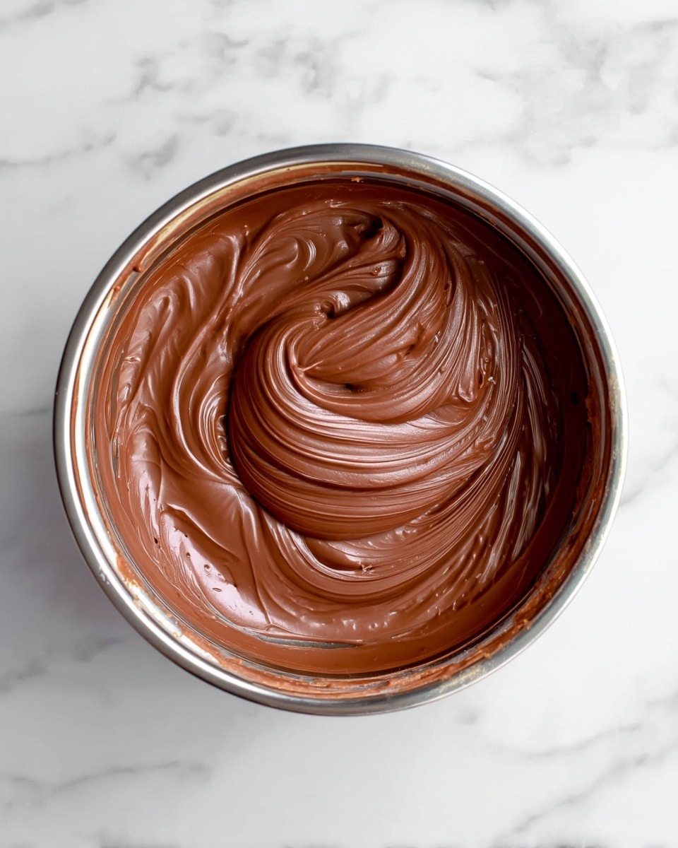 The image shows a close-up view of a silver bowl filled with smooth, creamy chocolate batter. The chocolate has a thick texture with shiny, swirling patterns on the surface, giving it a rich, glossy look. The batter fills most of the bowl, and the swirls create a wave-like design in the center, with light reflecting softly on the chocolate curves. The bowl is placed on a white marbled surface, and the photo is taken from above, focusing on the texture and color of the chocolate mixture. photo taken with an iphone --ar 4:5 --v 7
