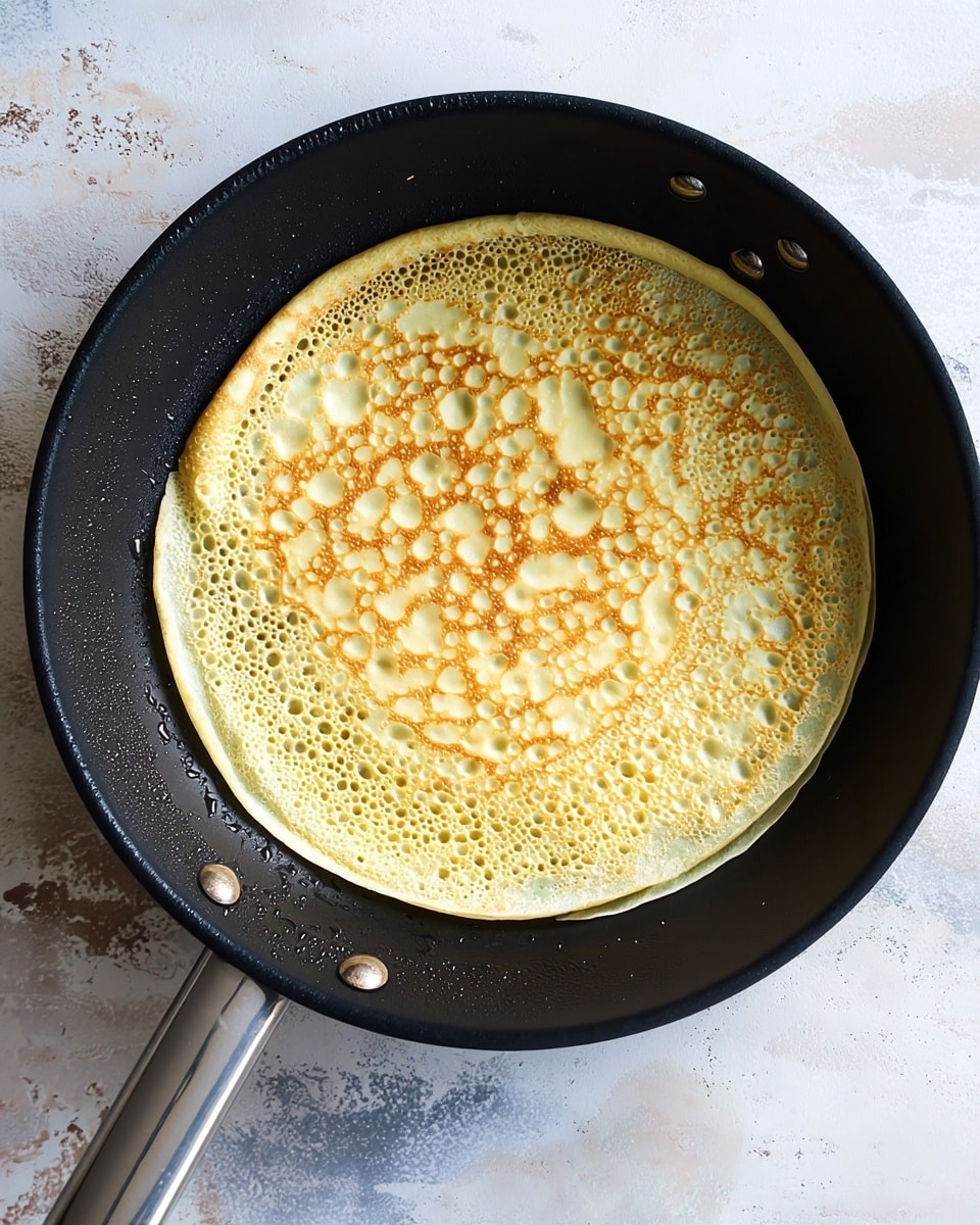 A single light yellow crepe with small air bubbles and a lightly browned pattern is cooking in a black frying pan with a silver handle, set on a white marbled surface. The crepe is thin and round, covering the whole pan base. The pan’s texture contrasts with the smooth and delicate crepe surface photo taken with an iphone --ar 4:5 --v 7