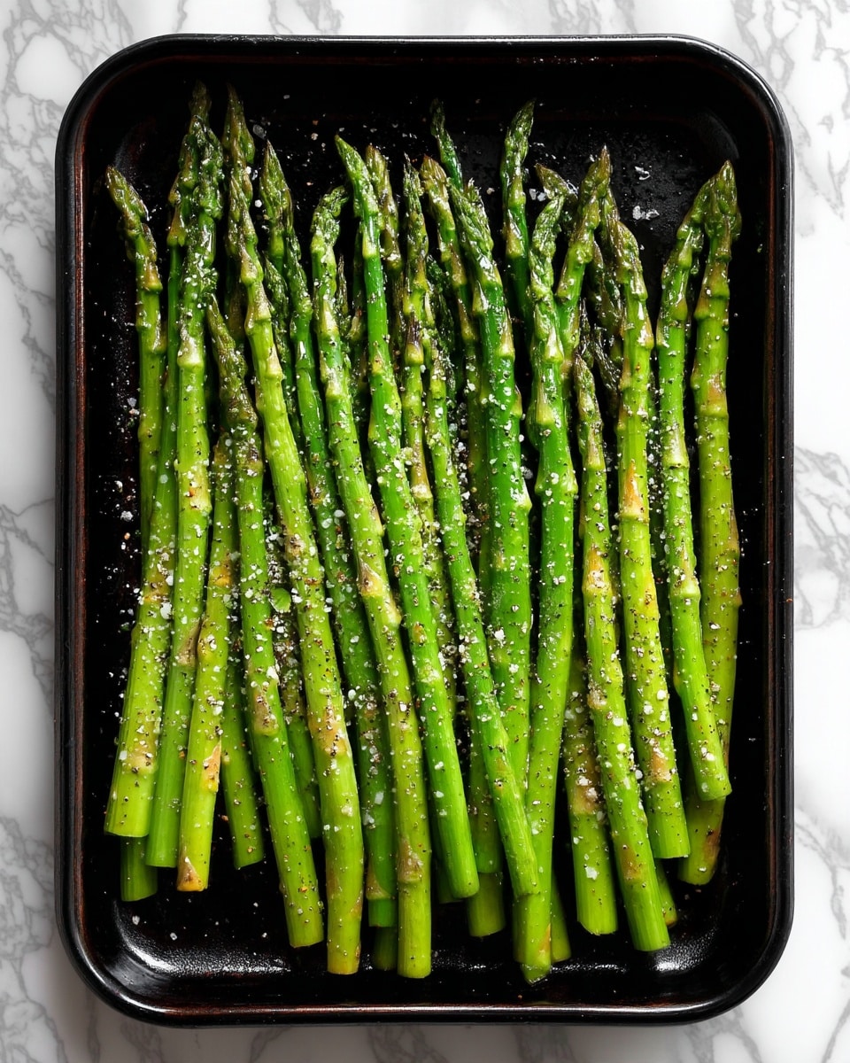 A black baking tray filled with a single layer of fresh green asparagus spears, neatly arranged side by side. The asparagus have a slight shine from olive oil and are sprinkled with coarse salt and black pepper, showing some light texture from the seasoning. The tips are tightly closed and the stalks are slender, with their natural green color contrasting against the dark tray. The background is a white marbled surface, visible around the edges of the tray. photo taken with an iphone --ar 4:5 --v 7
