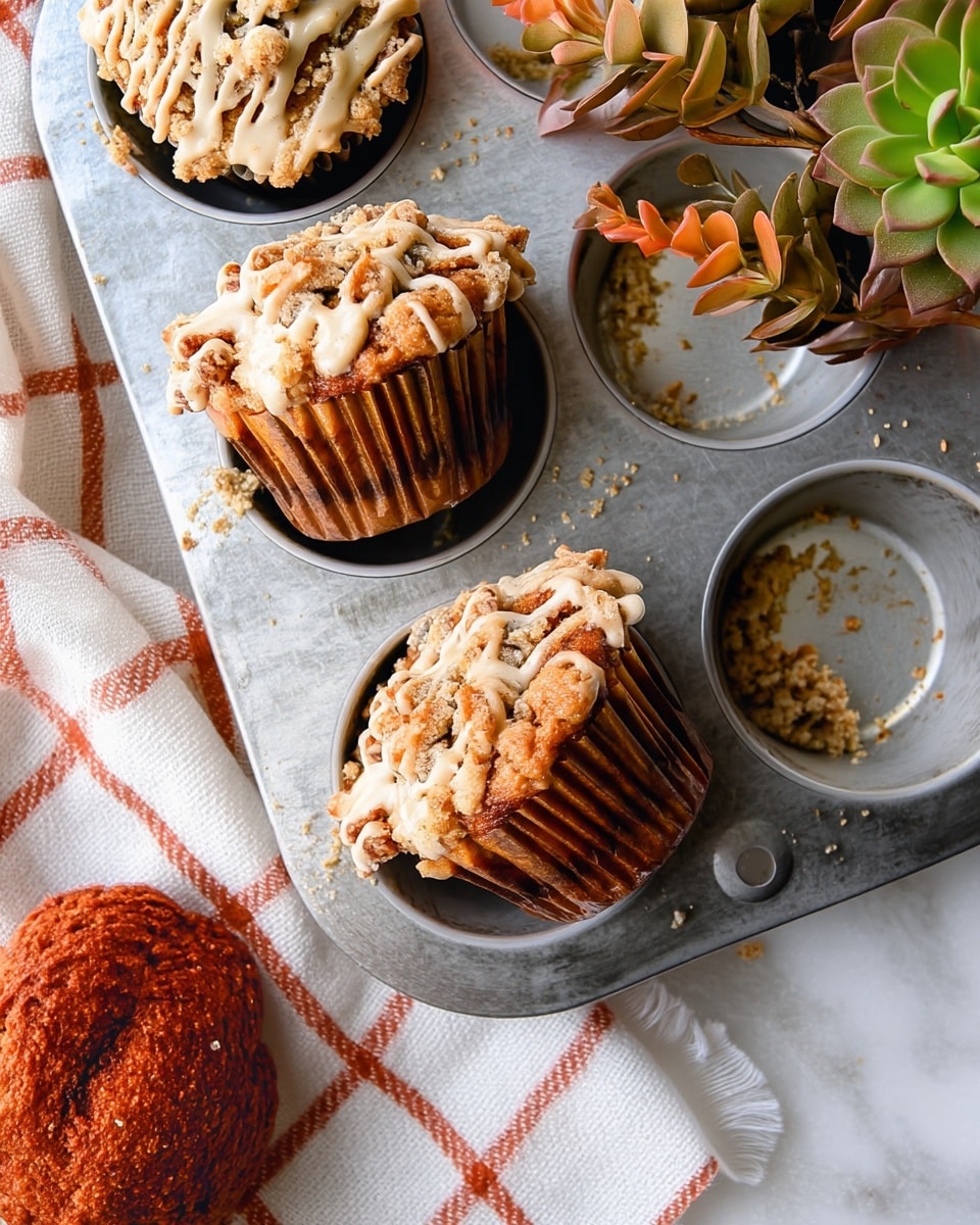 There are four muffins inside a metal muffin tray on a white marbled surface with an orange and cream checkered cloth nearby. Each muffin has two visible layers: a light golden brown crumbly top layer with a rough texture covered in drizzled creamy white icing, and a darker golden brown base layer with a smooth texture inside a brown paper muffin liner on one muffin. One slot in the tray is empty with crumbs around it, and there are some orange and green succulents in the top right corner. Photo taken with an iphone --ar 4:5 --v 7