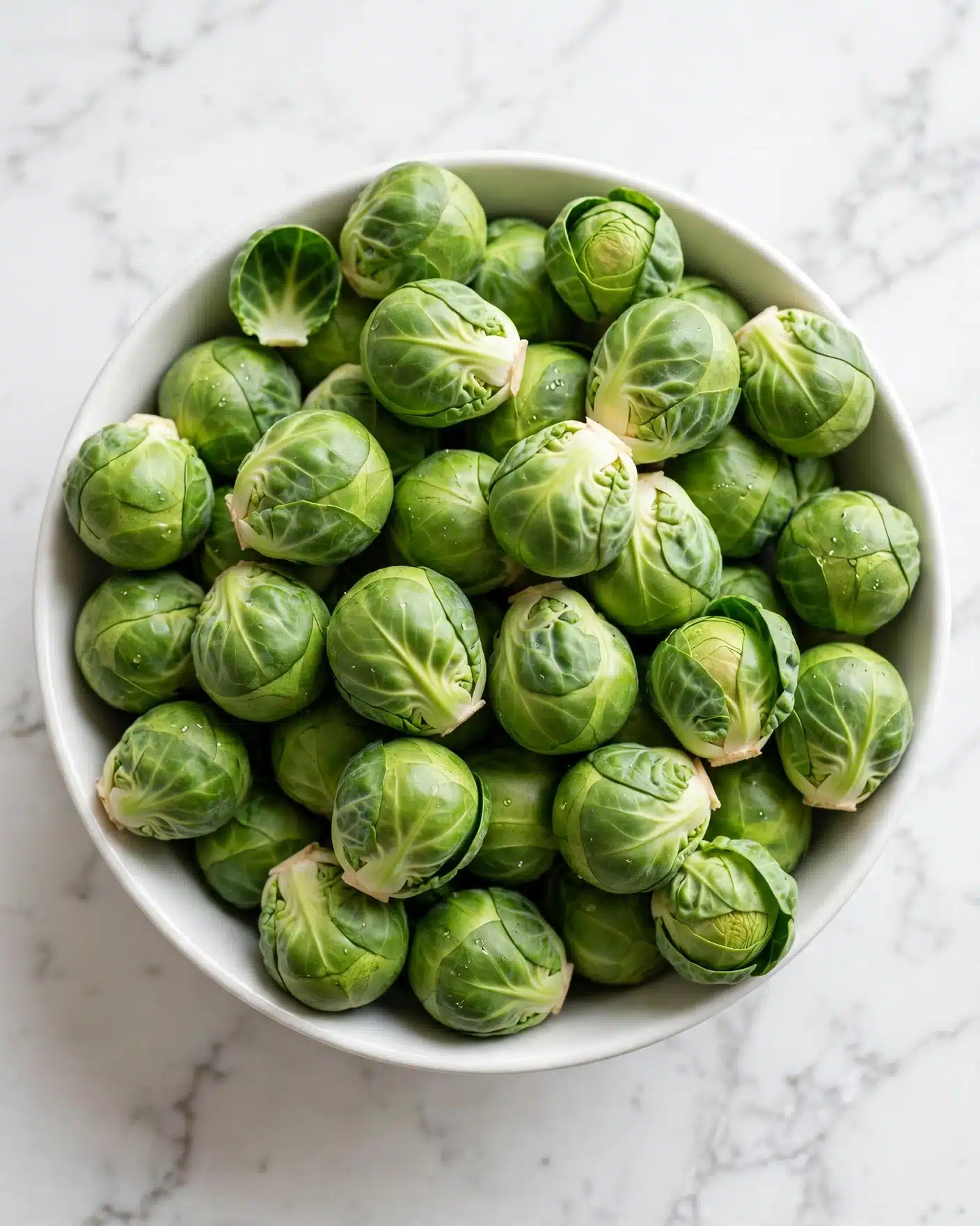 Bang Bang Brussels Sprouts Recipe 4 A white bowl filled with many fresh Brussels sprouts, each bright green with smooth, tightly layered leaves showing light and dark green shades. The bowl sits on a white marbled surface, adding clean contrast to the vibrant vegetables. The Brussels sprouts are piled evenly, some showing their round shape while others display their leafy texture in small clusters photo taken with an iphone --ar 4:5 --v 7