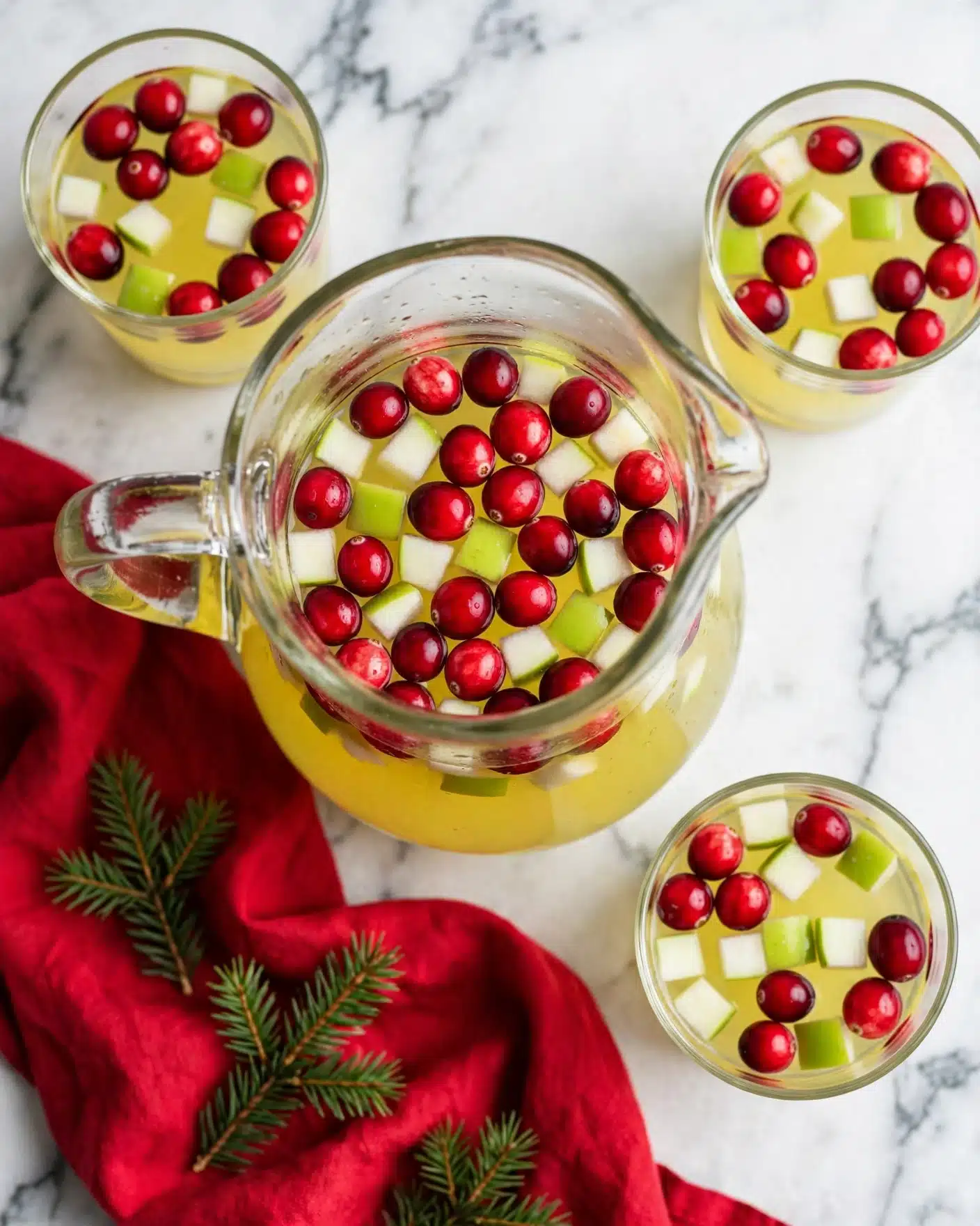 Sparkling Grape & Fruit Punch Recipe 5 A clear glass pitcher filled with a light yellow liquid containing many bright red cranberries and green apple cubes, all floating evenly. Surrounding the pitcher are three clear glasses each filled with the same yellow liquid and floating cranberries and green apple cubes, visible from above. A red cloth with soft folds lies next to the pitcher, partially on a white marbled surface, with some small sprigs of green pine resting on the cloth. The overall colors are red, green, and yellow with a fresh and festive feel. Photo taken with an iphone --ar 4:5 --v 7