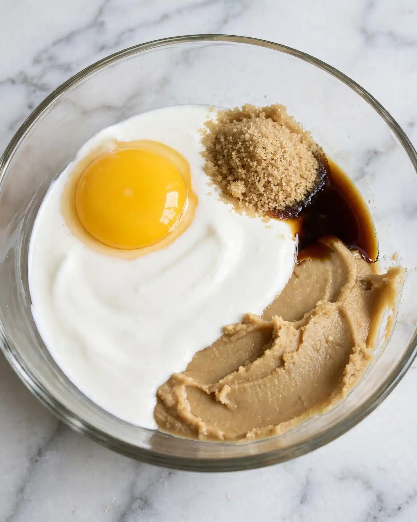 A clear glass bowl sits on a white marbled surface, containing separated ingredients in four distinct sections. On the left side, there is thick white cream forming a smooth base curving around the inside of the bowl. To the right of the cream is a light brown chunky layer with a slightly grainy texture. Above it, a rich dark brown syrup pools with a glossy surface. Centered over the cream, a whole raw egg yolk shines bright yellow with a smooth, round dome shape. Next to the yolk, a small pile of fine brown sugar with some darker spots rests on the cream. The ingredients are unmixed, showing clear borders between each layer photo taken with an iphone --ar 4:5 --v 7