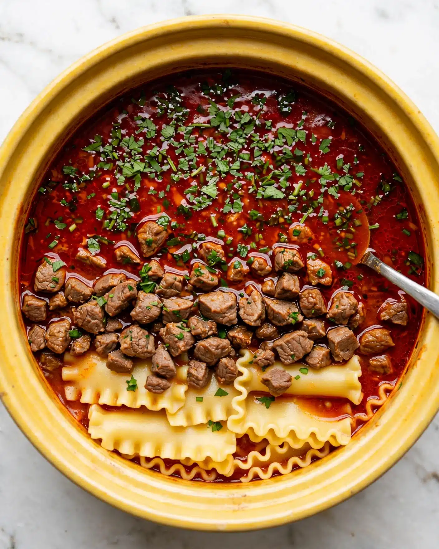 Easy Lasagna Soup Recipe 5 The image shows a large round yellow pot filled with a rich red soup. The soup has three layers of visible content: the top layer is a shiny red broth with green chopped herbs scattered over it, the middle layer contains small browned meat pieces and short curly pasta in light yellow, and the bottom layer reveals large flat pasta pieces with wavy edges in pale yellow. A silver spoon rests inside the pot, partly submerged in the broth. The pot sits on a white marbled surface. Photo taken with an iphone --ar 4:5 --v 7