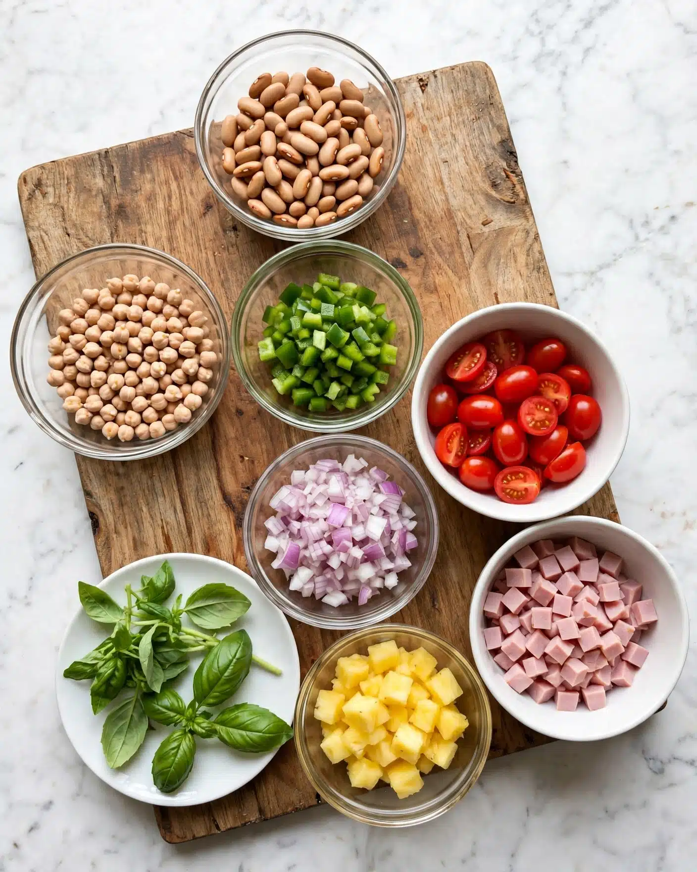 Italian Grinder Bean Salad Recipe 4 The image shows a wooden board with five clear glass bowls and two white bowls placed on it. One glass bowl at the top has light tan-colored beans, another glass bowl has small round beige chickpeas, and a third bowl contains small chopped green bell peppers. There is a white bowl filled with halved bright red cherry tomatoes and a glass bowl holding chopped light purple onions. Next to the onions, another white bowl contains small diced pieces of light pink ham. A white plate holds fresh green basil leaves and another glass bowl contains small yellow pineapple pieces. The background is a white marbled surface. photo taken with an iphone --ar 4:5 --v 7