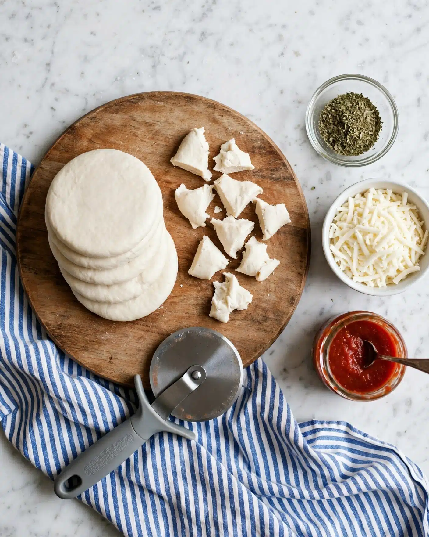 A round wooden board is placed on a white marbled surface with a blue and white striped cloth nearby. On the board, there are several thick white dough discs arranged in a small pile on the left side. To the right, there are small white dough pieces cut into irregular shapes. Below the board is a pizza cutter with a gray handle resting slightly on the cloth and board. Next to the board, on the right, are three bowls: a small clear glass bowl with green dried herbs at the top, a white bowl filled with shredded white cheese below it, and a small glass jar of red tomato sauce with a small spoon inside at the bottom. Photo taken with an iphone --ar 4:5 --v 7
