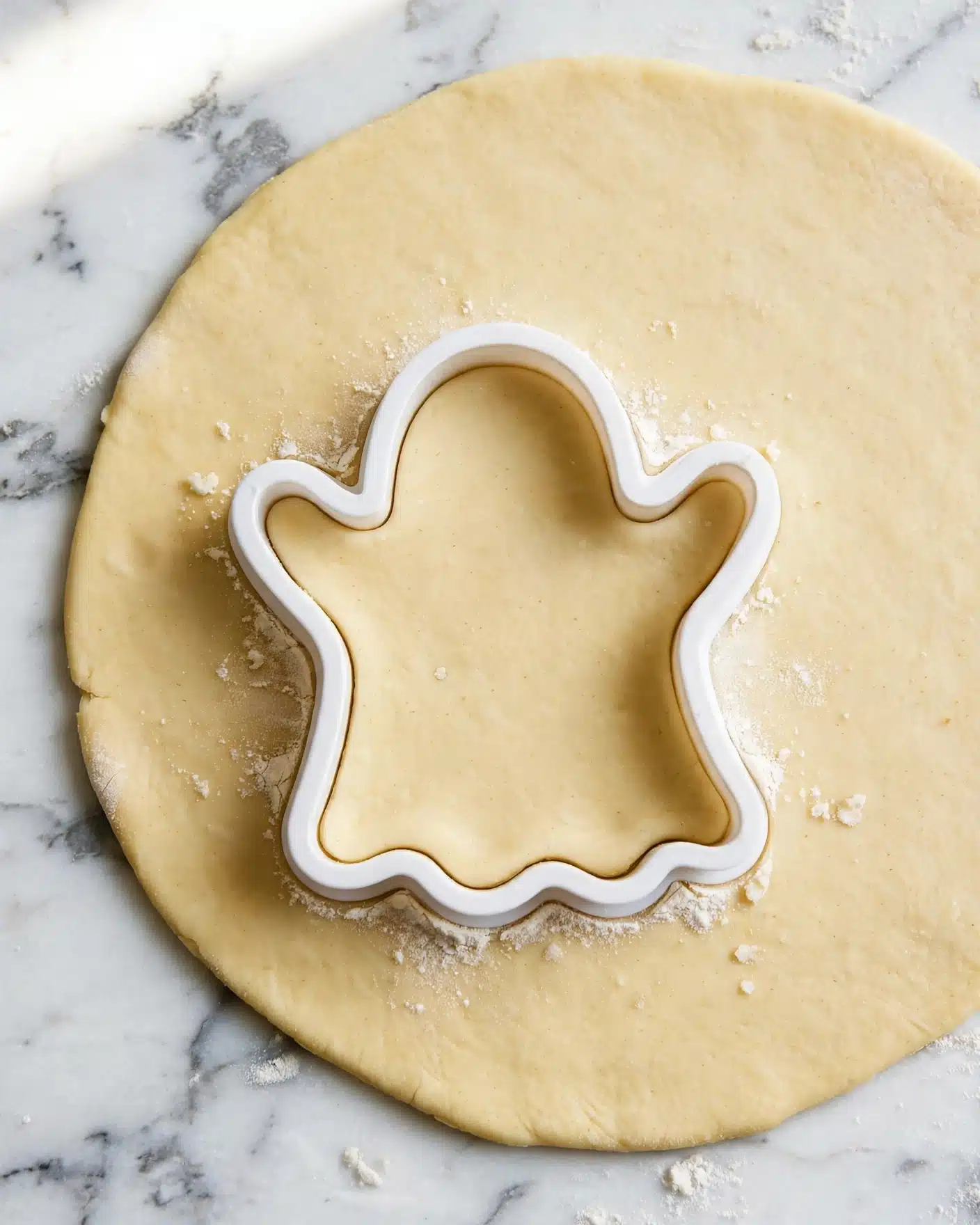 A flat, light yellow dough sheet is laid out on a white marbled surface. On top of the dough, there is a white cookie cutter shaped like a ghost, pressed into the dough, forming a clear ghost shape with rounded edges and two raised arms. The dough inside the cutter is still connected to the rest, showing a smooth texture with a slight dusting of flour around the cutter's edge. photo taken with an iphone --ar 4:5 --v 7