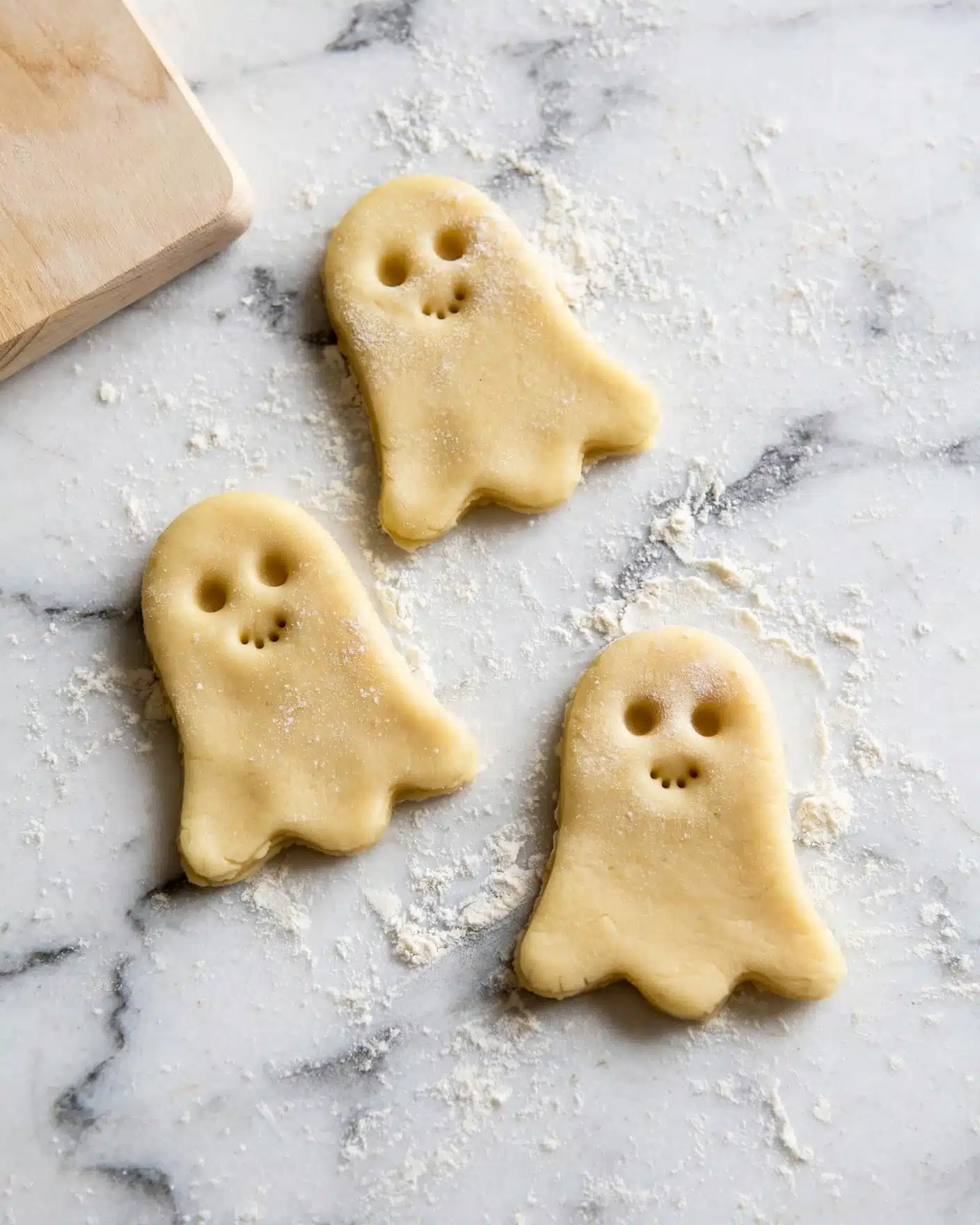 Three ghost-shaped cookie dough pieces lie flat on a white marbled surface lightly dusted with flour. Each ghost has small round eyes and simple mouth details made by small indentations in the dough. The dough is pale yellow and has a smooth texture with a slight dusting of flour on top. A light-colored wooden cutting board is partially visible in the top left corner. photo taken with an iphone --ar 4:5 --v 7