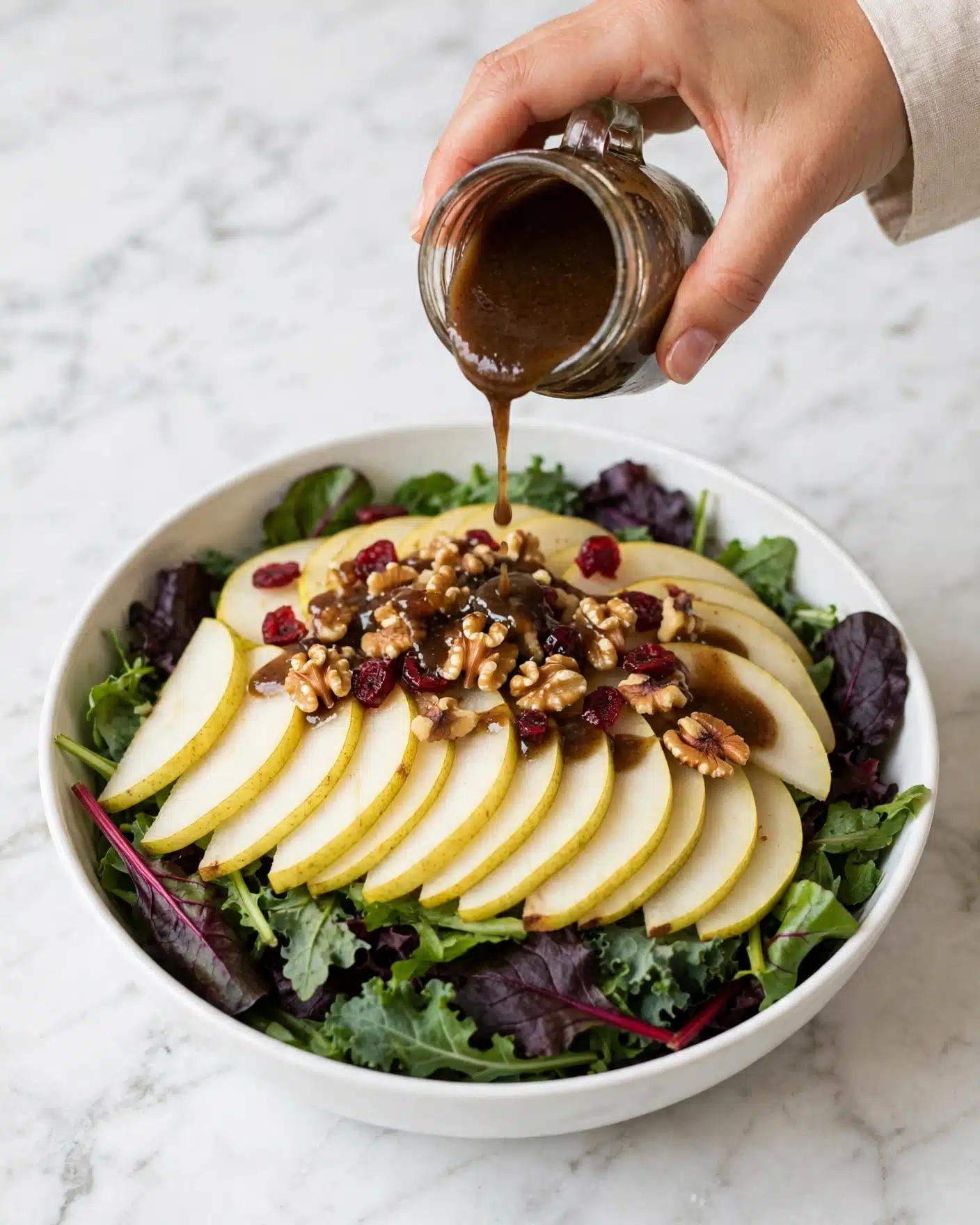 Autumn Pear Salad with Candied Walnuts and Balsamic Vinaigrette Recipe 5 A white bowl holds a fresh salad with three main layers: the bottom layer is a mix of dark green and purple leafy greens, the middle layer has light yellow pear slices arranged all over, and the top layer is scattered with small brown walnut pieces and red dried cranberries. A woman's hand is pouring a dark brown dressing over the salad. The background surface is a white marbled texture. photo taken with an iphone --ar 4:5 --v 7