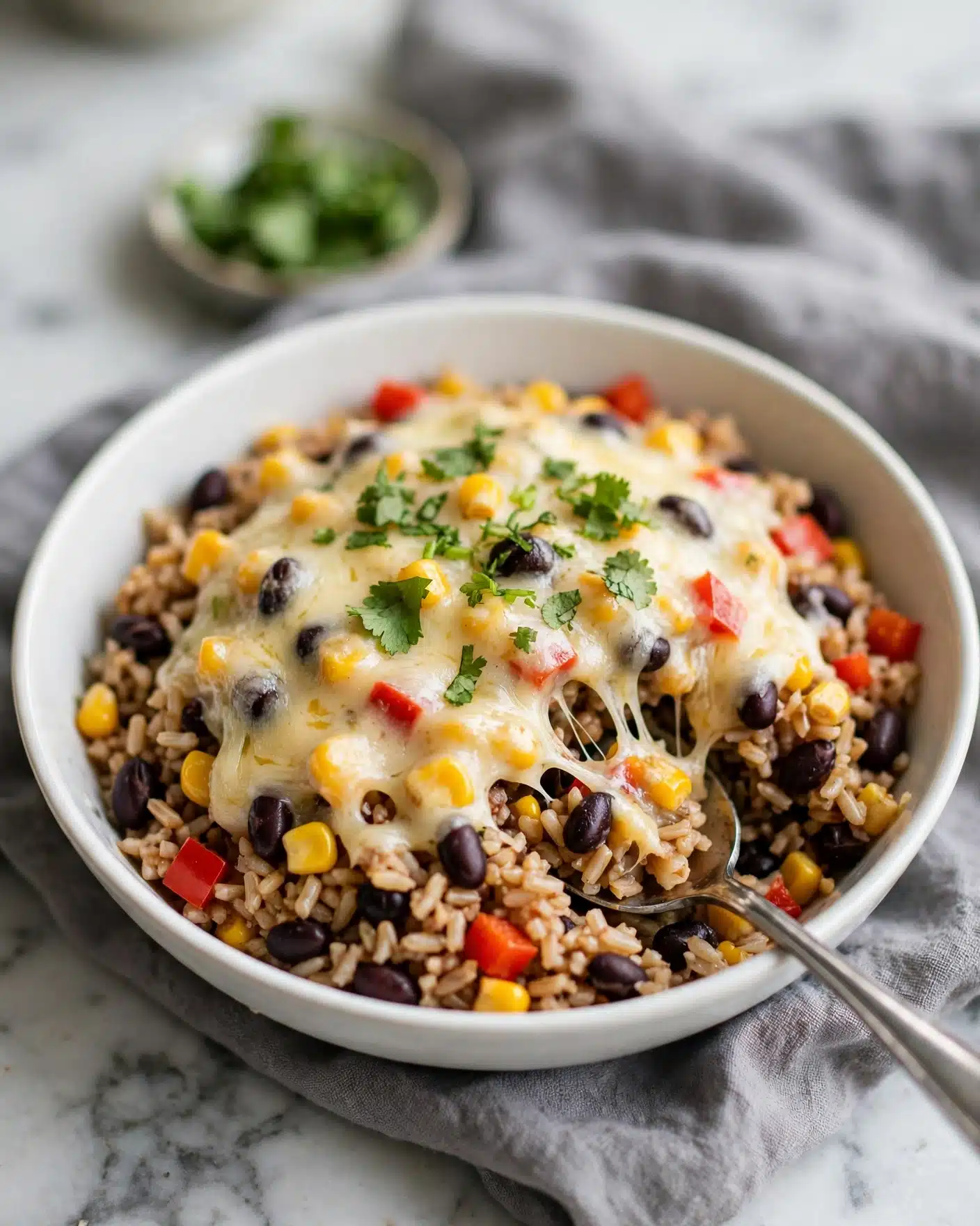Mexican Ground Beef Casserole with Rice Recipe 5 In a white bowl, there is a mixed dish with two main layers: the bottom layer is a mix of brown rice with small pieces of red bell pepper, black beans, and yellow corn kernels, giving it a colorful and textured look; the top layer has melted light yellow cheese that stretches slightly, mixed with some more corn, beans, and red pepper pieces; fresh green herbs are sprinkled on top for a bright touch; a silver spoon is scooping into the bowl from the bottom right, resting inside the bowl; the bowl is placed on a gray cloth with a blurred small dish of green garnish in the background, all sitting on a white marbled surface photo taken with an iphone --ar 4:5 --v 7