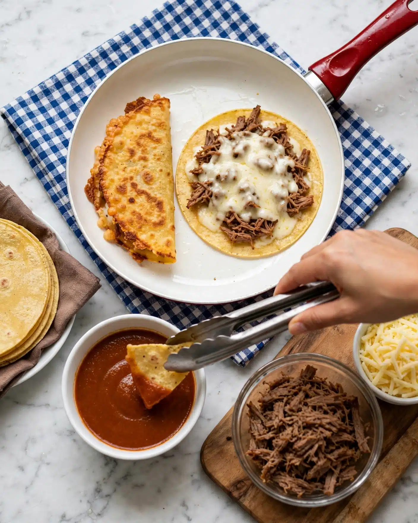 Slow-Cooker Birria Tacos Recipe 4 The image shows a food preparation scene featuring two soft corn tortillas with a golden yellow color on a white frying pan with a red handle; one tortilla is folded and fried with a crunchy texture, while the other is open and topped with shredded brown meat and melted cheese in a creamy white color. Nearby, a white bowl holds a reddish brown sauce with a smooth texture, with a woman's hand using tongs to dip a tortilla chip into the sauce. On a wooden board, a glass bowl contains shredded brown meat pieces, and a small white bowl holds shredded pale yellow cheese. To the side, there is a white plate lined with brown cloth holding more soft yellow tortillas. The scene is set on a white marbled surface with a checkered cloth under the frying pan. Photo taken with an iphone --ar 4:5 --v 7