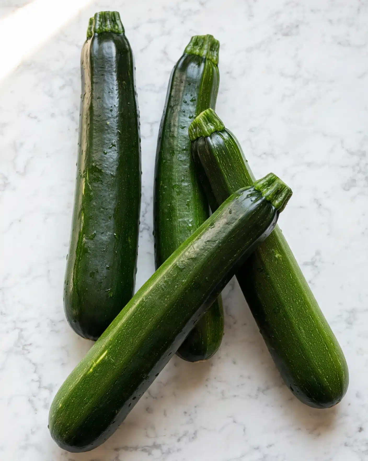 Easy Quick and Easy Baked Zucchini Recipe 4 The image shows four whole zucchinis laid out on a white marbled surface. Each zucchini is dark green with smooth, shiny skin and small lighter green streaks, with the stem end on top. They are arranged in a loose cluster, with two zucchinis standing more upright and the other two leaning slightly. The light reflects softly on their surfaces, showing their fresh texture. photo taken with an iphone --ar 4:5 --v 7