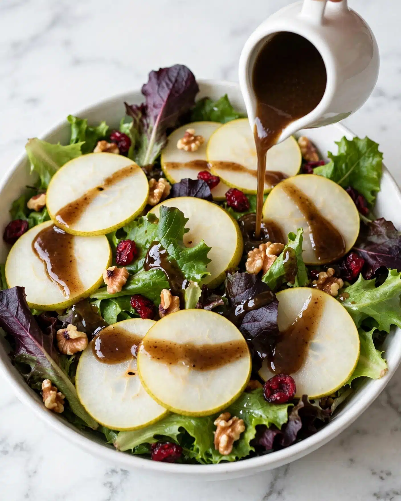 The dish is a fresh salad in a white bowl on a white marbled surface. The bottom layer is a mix of green and purple leafy salad leaves with a fresh texture. Over these are thin, round, light yellow slices of pear arranged all over the salad. There are small pieces of brown walnuts scattered among the leaves and pear slices, adding texture. Bright red dried cranberries are also spread over the layers for color contrast. A dark brown dressing is being poured from a small white sauce jug onto the salad, creating a glossy shine on some leaves and nuts. Photo taken with an iphone --ar 4:5 --v 7