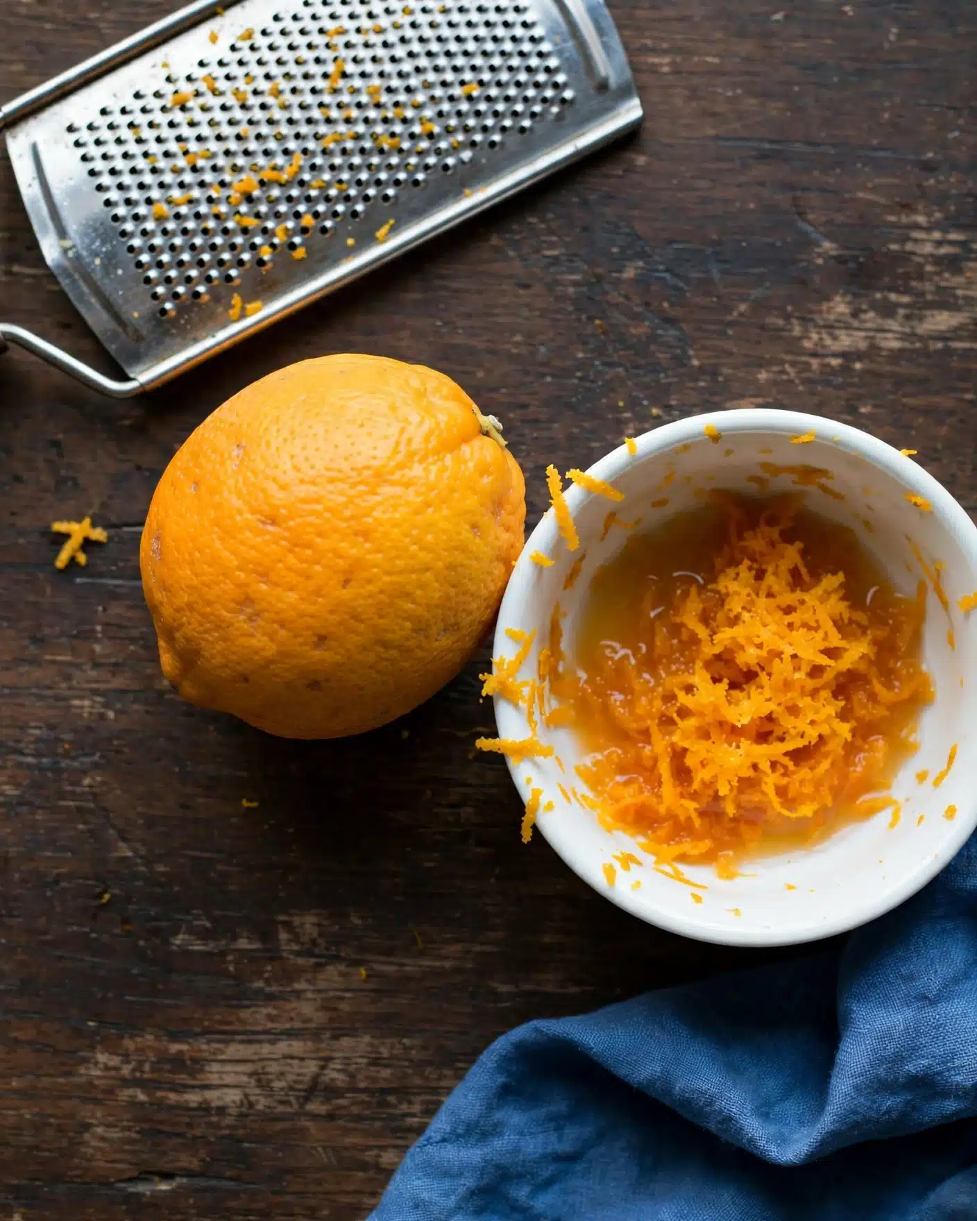 The image shows a round orange lemon with a textured peel placed on a dark wooden surface next to a small white bowl filled with grated orange zest and juice. At the top left, there is a metal grater with fine holes, and at the bottom right, part of a blue cloth is visible, resting on the same dark surface. The lemon is whole and positioned near the bowl, which has some zest stuck on its edge, showing the fresh citrus preparation. Photo taken with an iphone --ar 4:5 --v 7