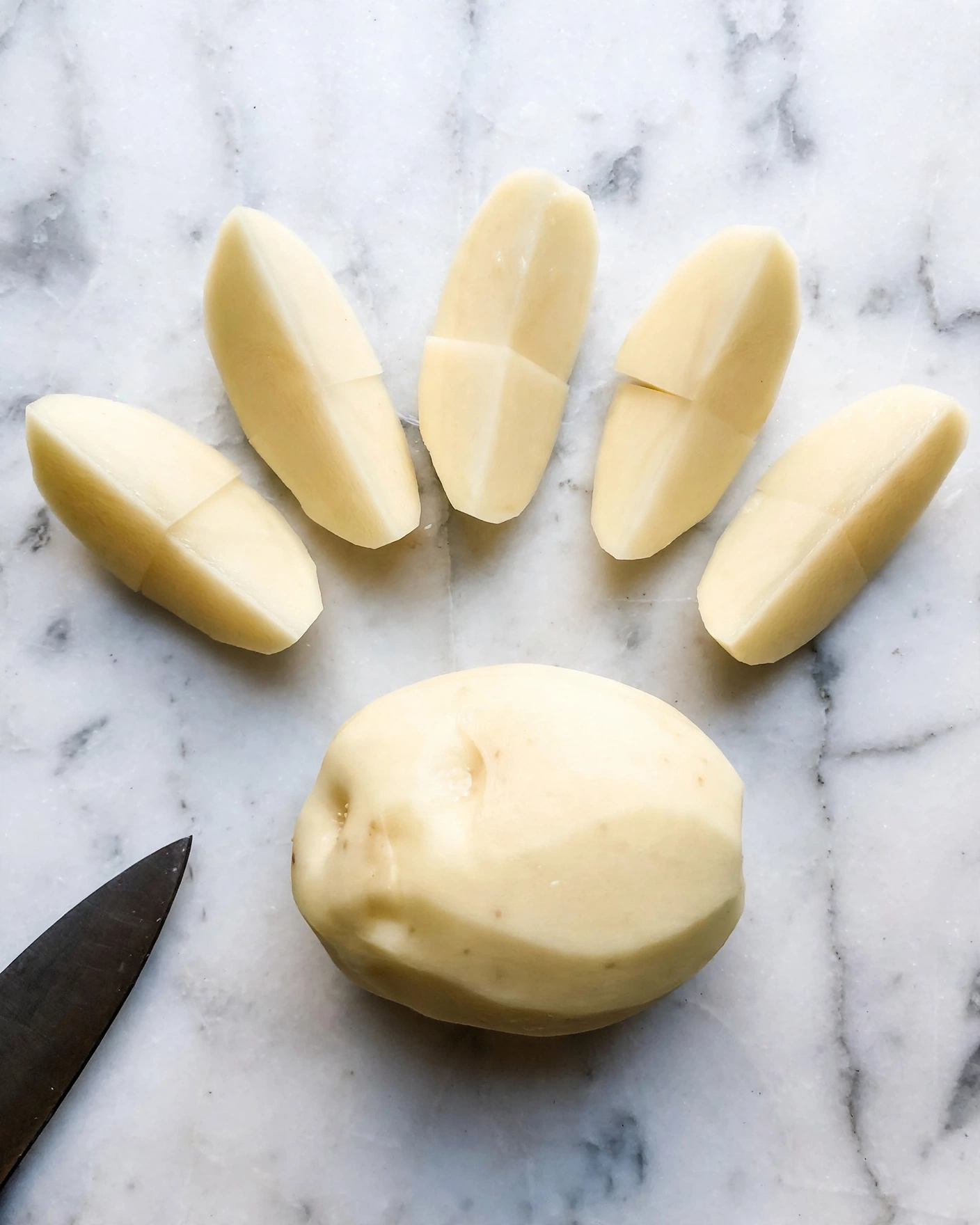 The image shows a peeled whole potato placed near the bottom center on a white marbled surface. Above it, there are five peeled potato wedges arranged loosely in a semi-circle, each wedge showing a smooth, soft yellowish-white texture. At the lower left corner of the image, part of a dark knife blade is visible, contrasting with the light potatoes and surface. The overall setting is bright and clean with natural light, showing simple preparation stages of the peeled potato pieces photo taken with an iphone --ar 4:5 --v 7