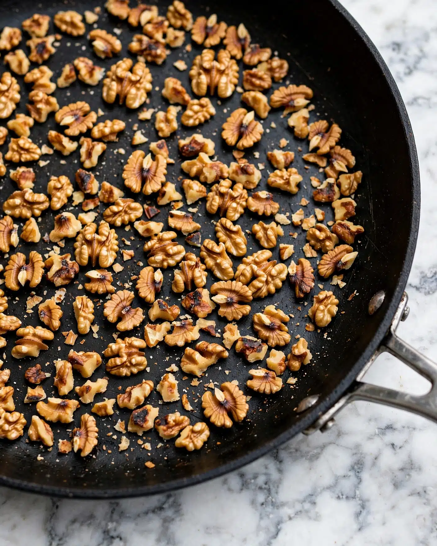 A close-up view of many small pieces of toasted walnuts scattered evenly in a black frying pan with a metal handle, showing a mix of light brown and golden brown colors with some darker caramelized spots. The pan is placed on a white marbled surface, and the walnuts have a rough, uneven texture with some broken parts. Photo taken with an iphone --ar 4:5 --v 7
