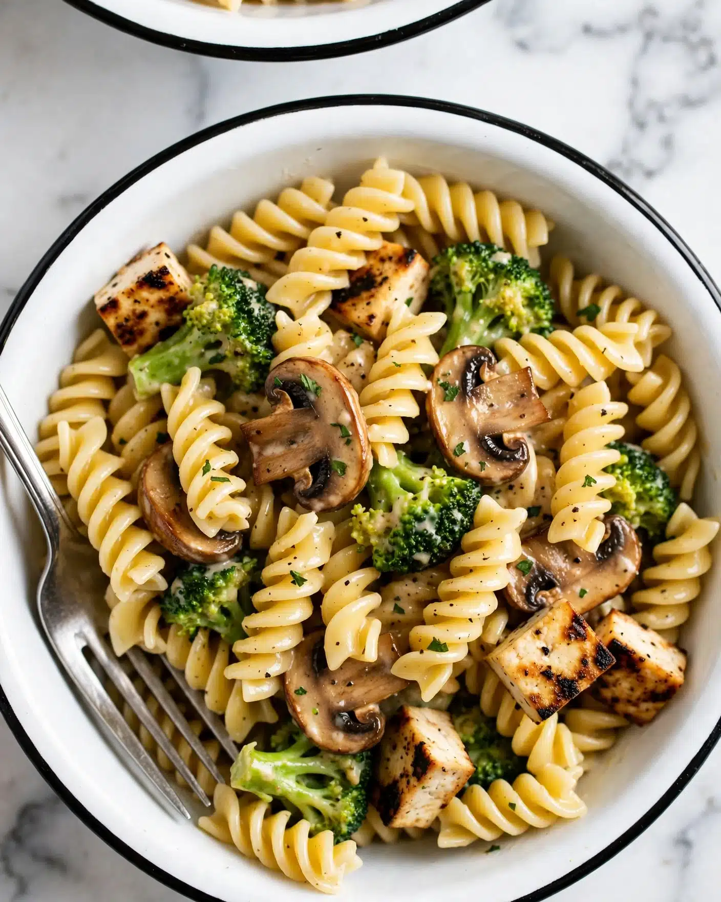 Healthier Broccoli Chicken Casserole Recipe 5 A white bowl with a black rim holds a close-up of mixed pasta made of spiral rotini, pale yellow in color, tossed with bright green broccoli florets and browned sliced mushrooms. A few small pieces of grilled tofu with a slight char are scattered in, all coated lightly with a creamy sauce. Black pepper and small herb bits are sprinkled on top. A silver fork rests inside the bowl on the left side, and part of another white bowl with black rim is visible at the top edge. The background is a white marbled texture. Photo taken with an iphone --ar 4:5 --v 7