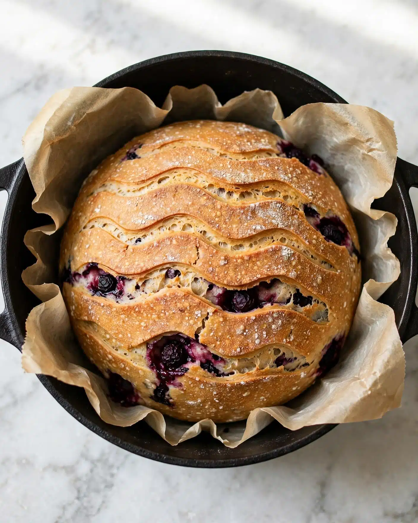 Lemon Blueberry Sourdough Bread Recipe 4 A round loaf of bread with a golden brown crust sits in a black pot lined with parchment paper. The bread has several dark purple spots inside, showing juicy berries baked into it. The top crust has wavy, scored lines creating a textured pattern, with small white specks of flour or sugar scattered lightly. The pot is positioned on a white marbled surface with a soft natural light shining from above. Photo taken with an iphone --ar 4:5 --v 7