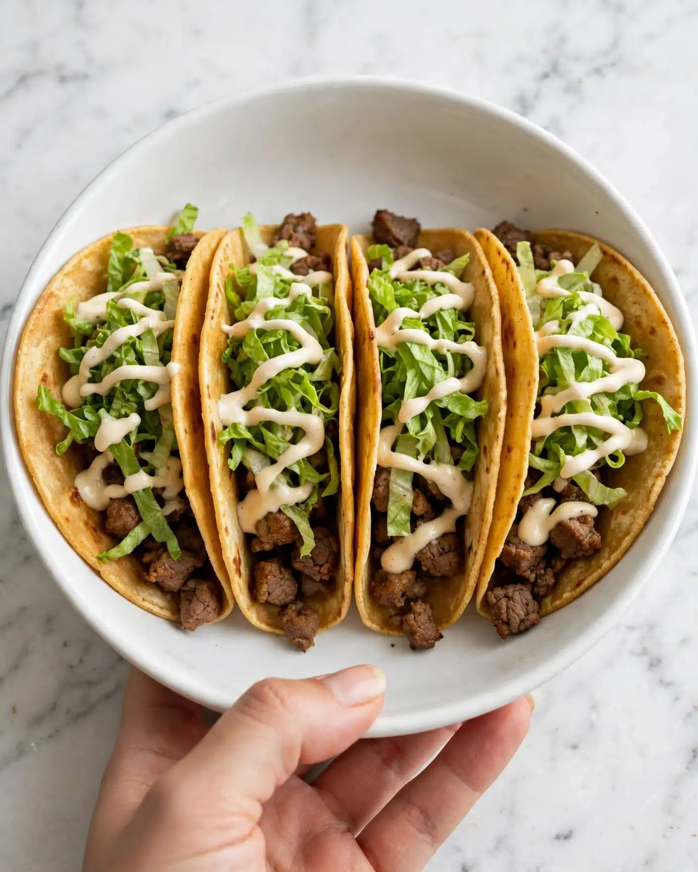 A white bowl holds four tacos arranged side by side, each taco filled with a base of small browned pieces of meat, topped with bright green shredded lettuce, and drizzled with a light cream sauce. The tacos have warm, golden-brown soft shells that are slightly folded. A woman's hand is holding the bowl from the bottom edge, set against a white marbled surface. photo taken with an iphone --ar 4:5 --v 7