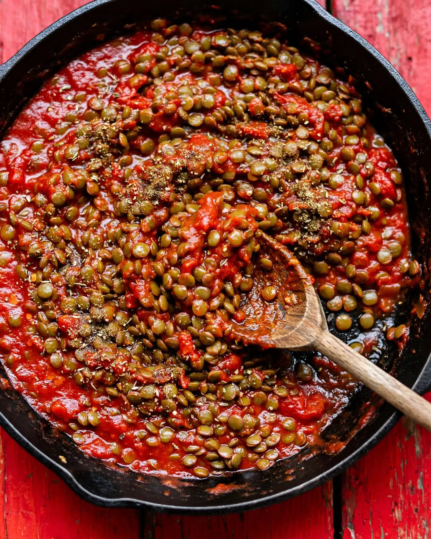 Vegan Sloppy Joes Recipe 4 The image shows a black cast iron pan filled with green lentils cooked in a thick red tomato sauce base. The lentils cover most of the pan's surface with some scattered seasoning powders on top, adding dark brown and beige specks over the green lentils and red sauce. A wooden spoon sits in the pan, touching the food, mixing the lentils and sauce. The pan sits on a bold red wooden surface, creating a strong color contrast. photo taken with an iphone --ar 4:5 --v 7